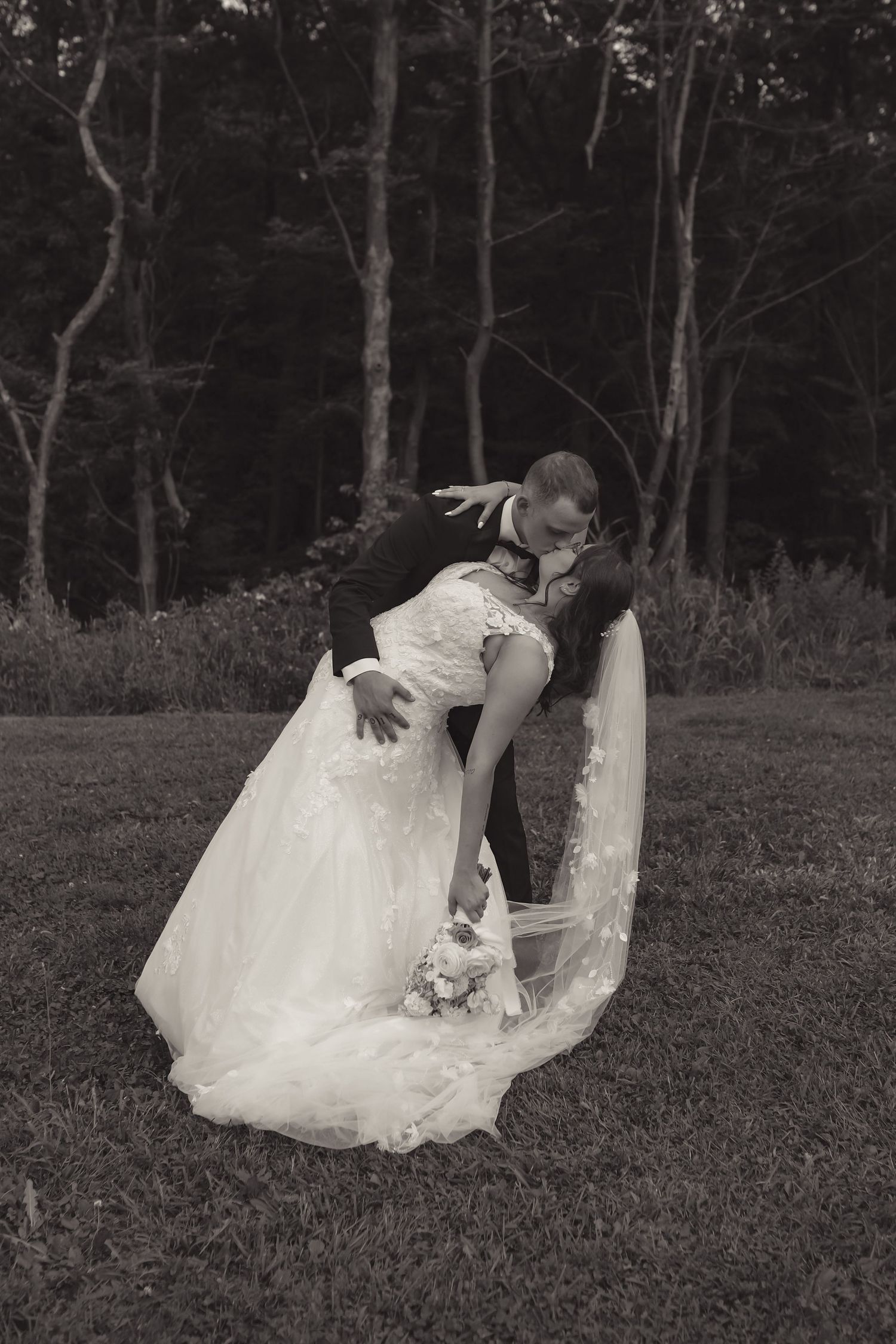 Bride and groom share romantic dip kiss outdoors in forest, wedding dress with veil, black and white.