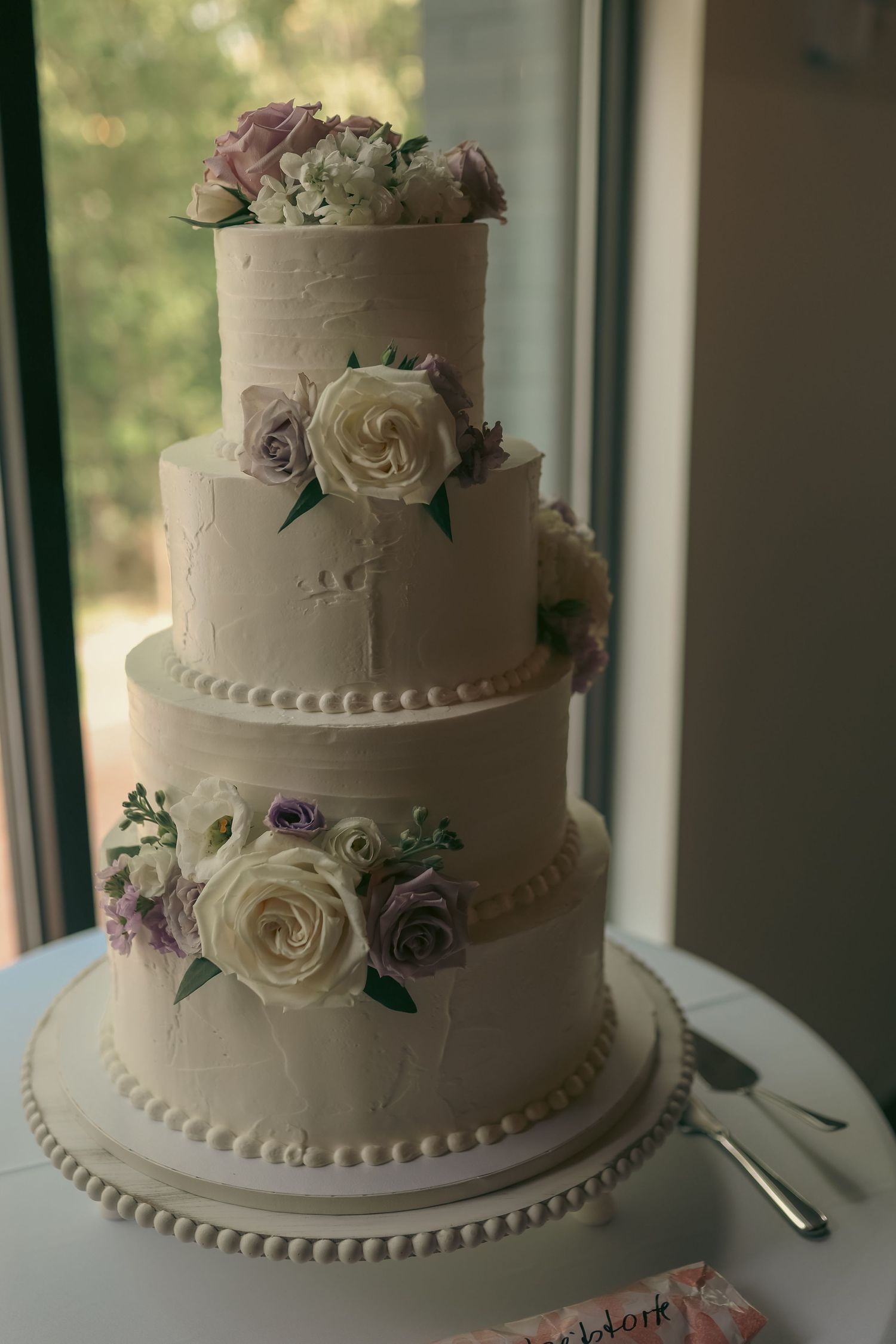 Elegant three-tier white wedding cake adorned with cream and lavender roses on a white cake stand.
