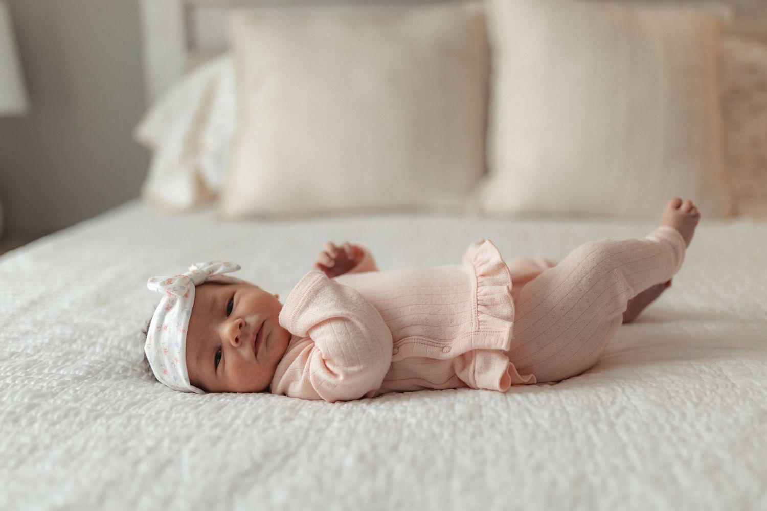Newborn baby in pink outfit and white bow headband lying on a white bed.