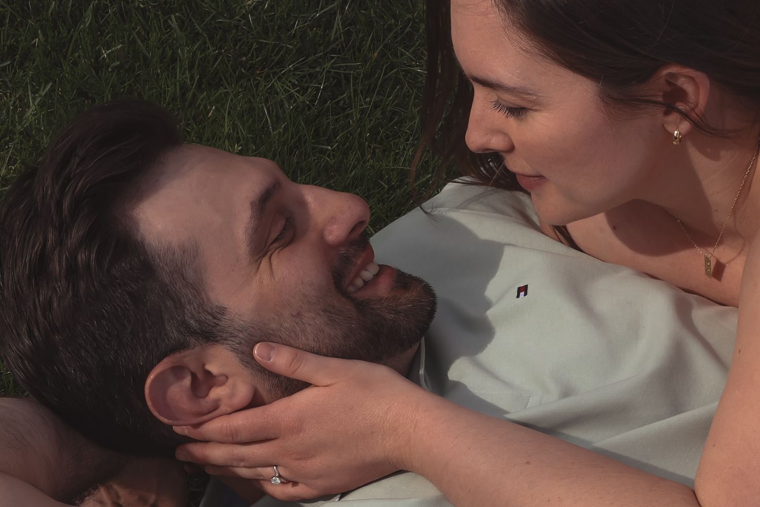 Romantic couple lying on grass, woman tenderly holding man's face, sharing an intimate moment outdoors.