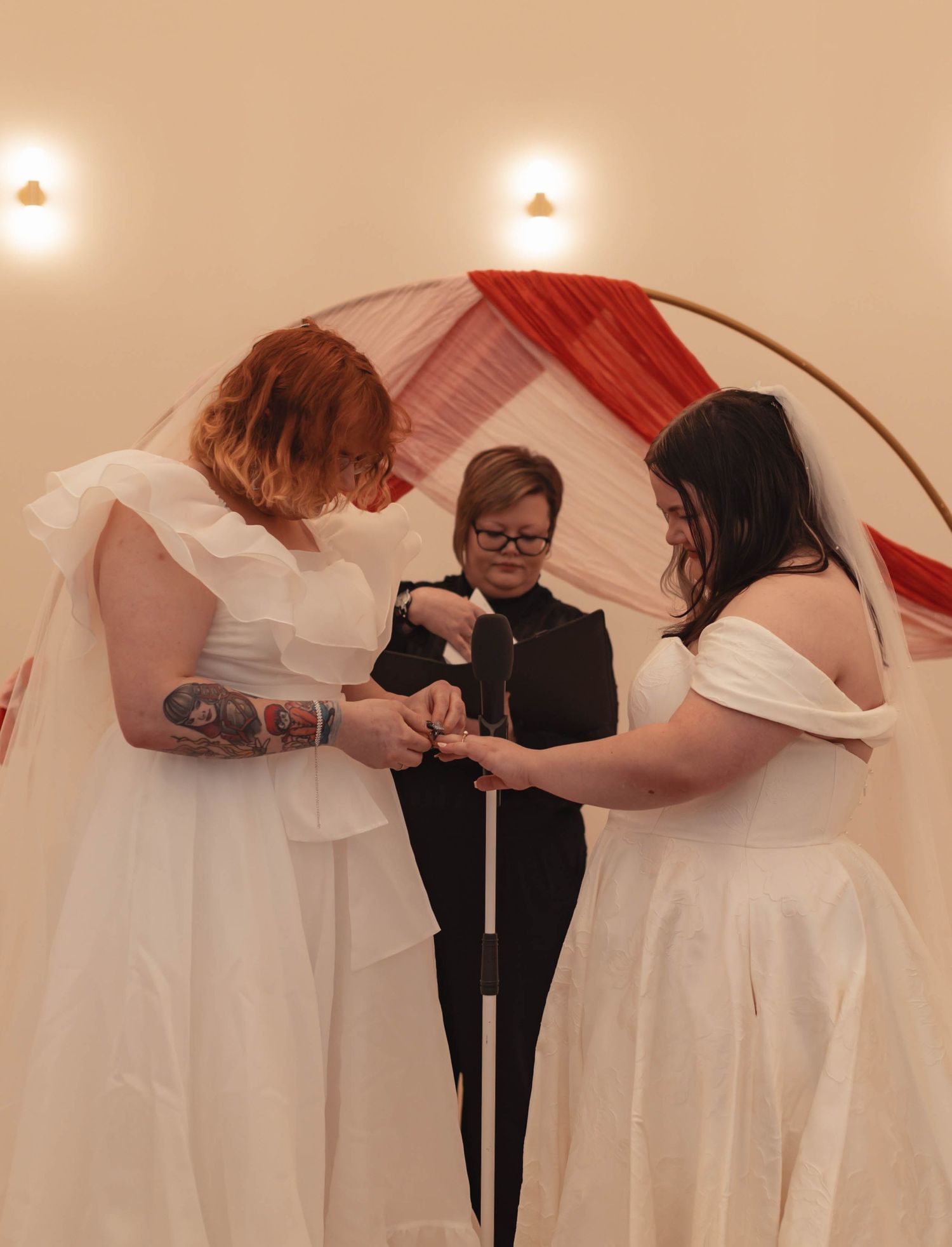 Two brides exchanging rings during wedding ceremony with officiant beneath a floral arch.