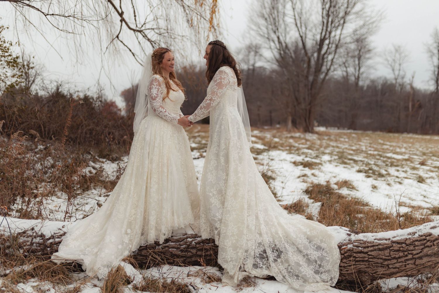 Two brides in white lace gowns hold hands on a snowy winter landscape, standing on a fallen log.