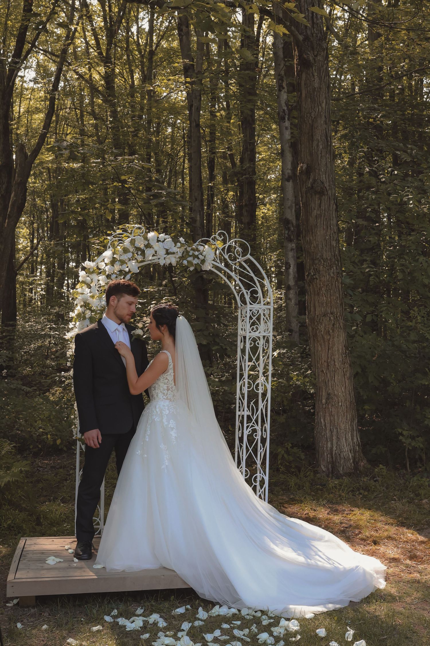 Bride and groom stand beneath white floral arch in sunlit forest wedding ceremony.