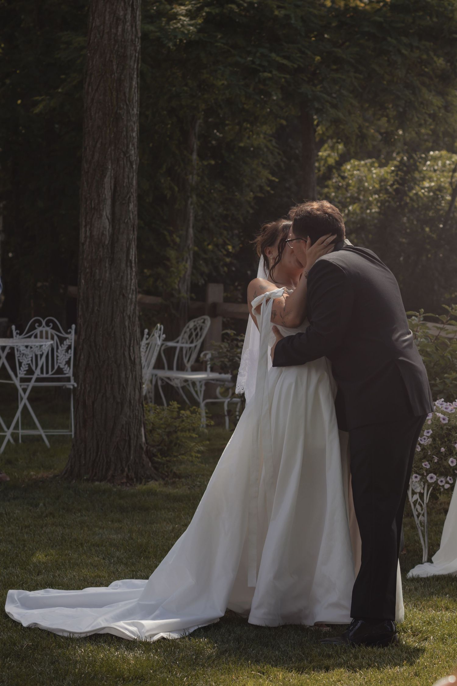A bride and groom share their first kiss outdoors in a sunlit garden during their wedding ceremony.