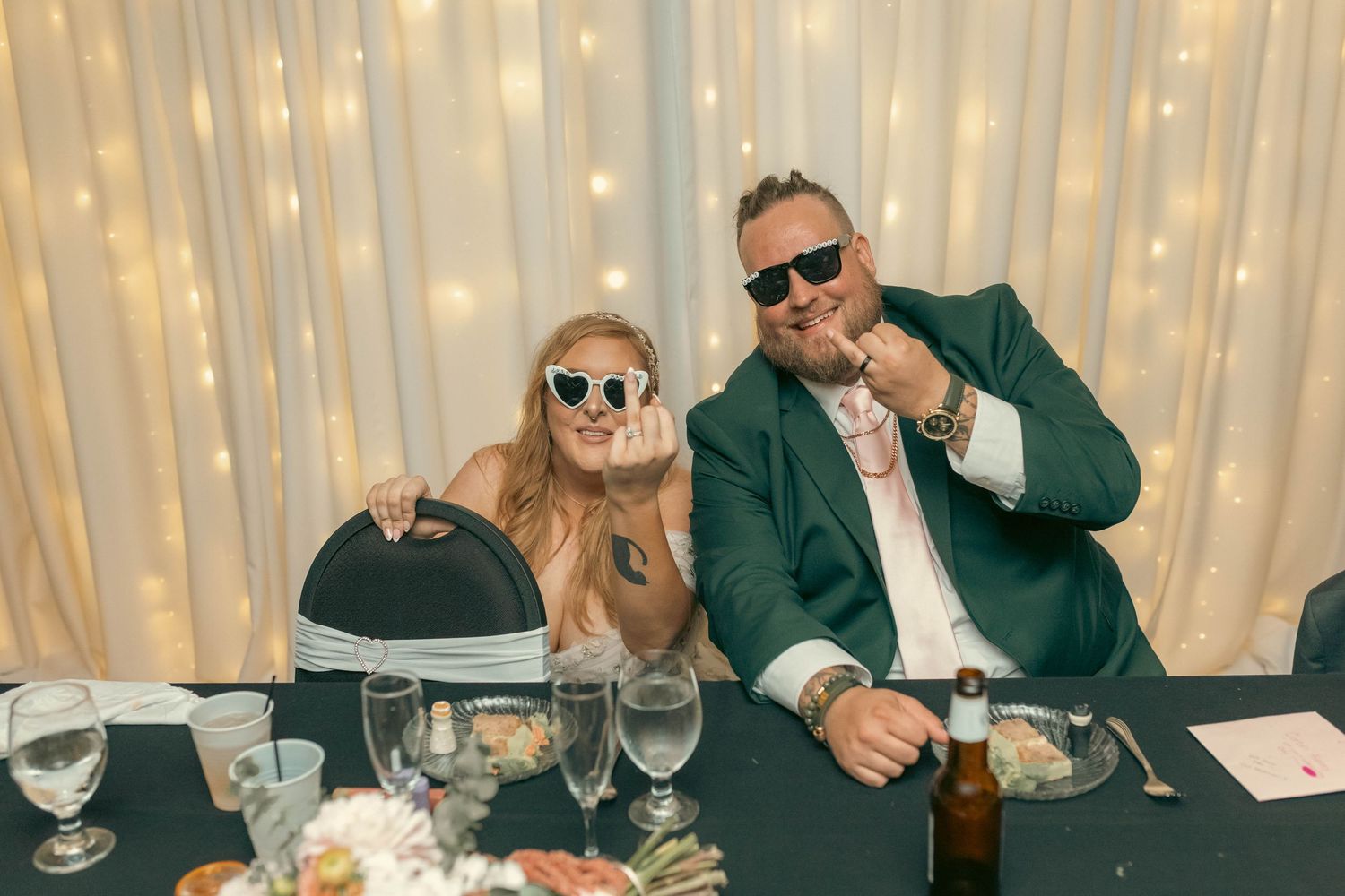 Two wedding guests wearing sunglasses pose playfully at a reception table with fairy light backdrop.
