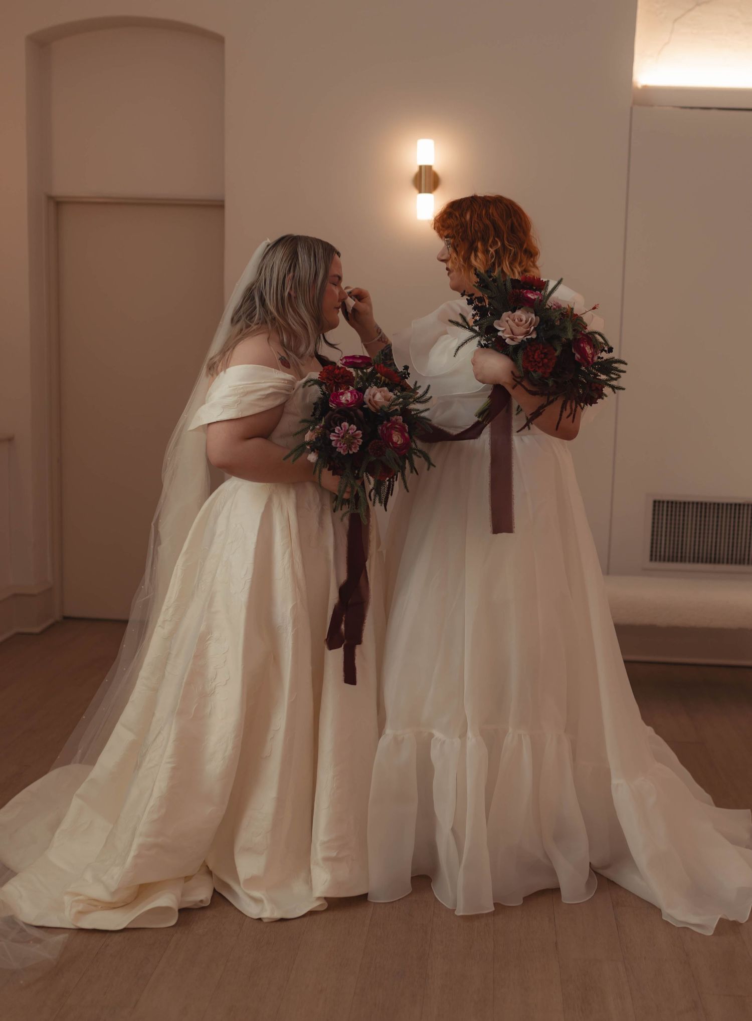 Two brides in white wedding gowns holding dark floral bouquets and sharing a tender moment together.