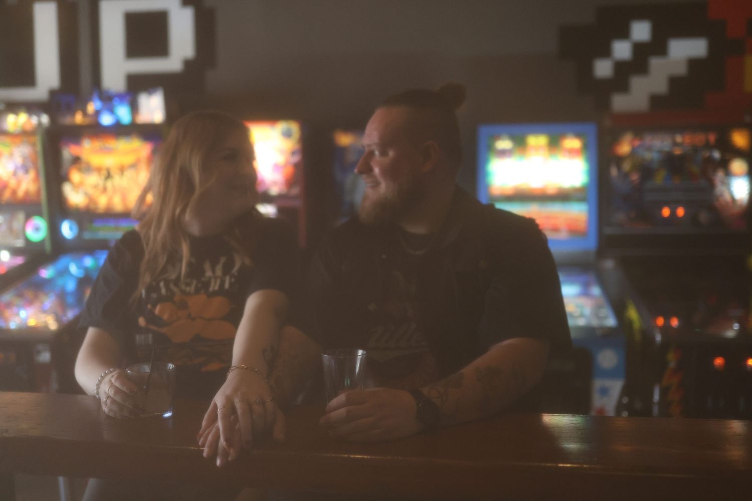 Couple sharing a romantic moment at a dimly lit arcade bar with colorful pinball machines in the background.