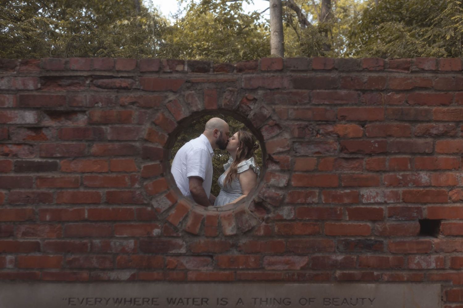 Couple sharing a romantic kiss framed through a circular opening in a rustic red brick garden wall.