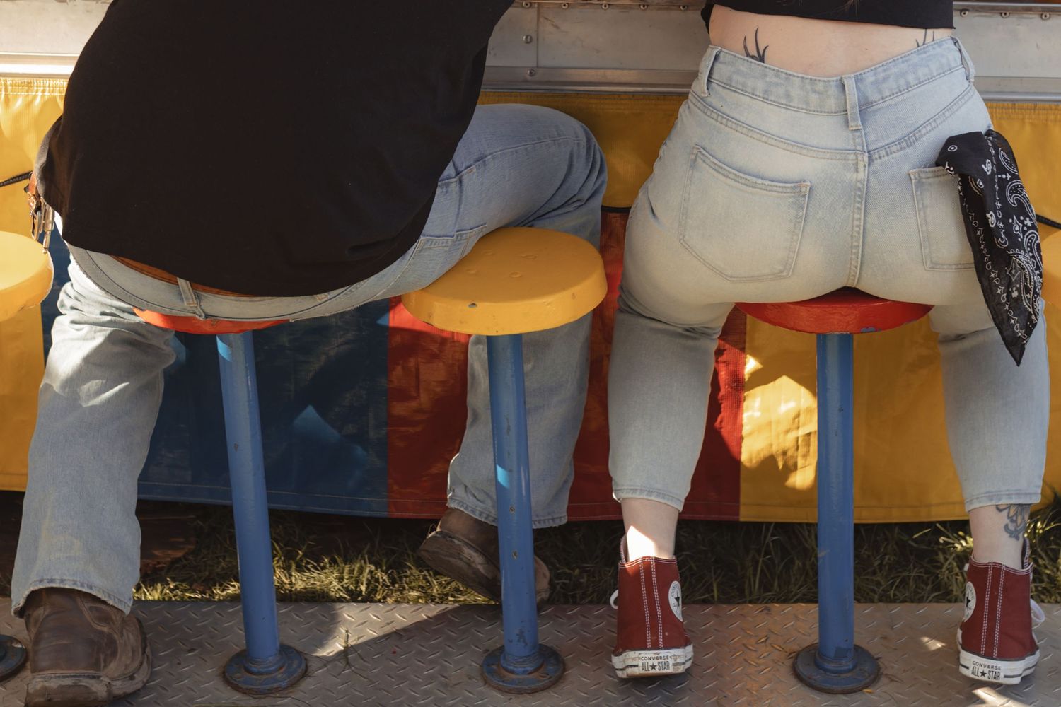 Two people sitting on colorful stools at an outdoor counter, wearing jeans and casual shoes.