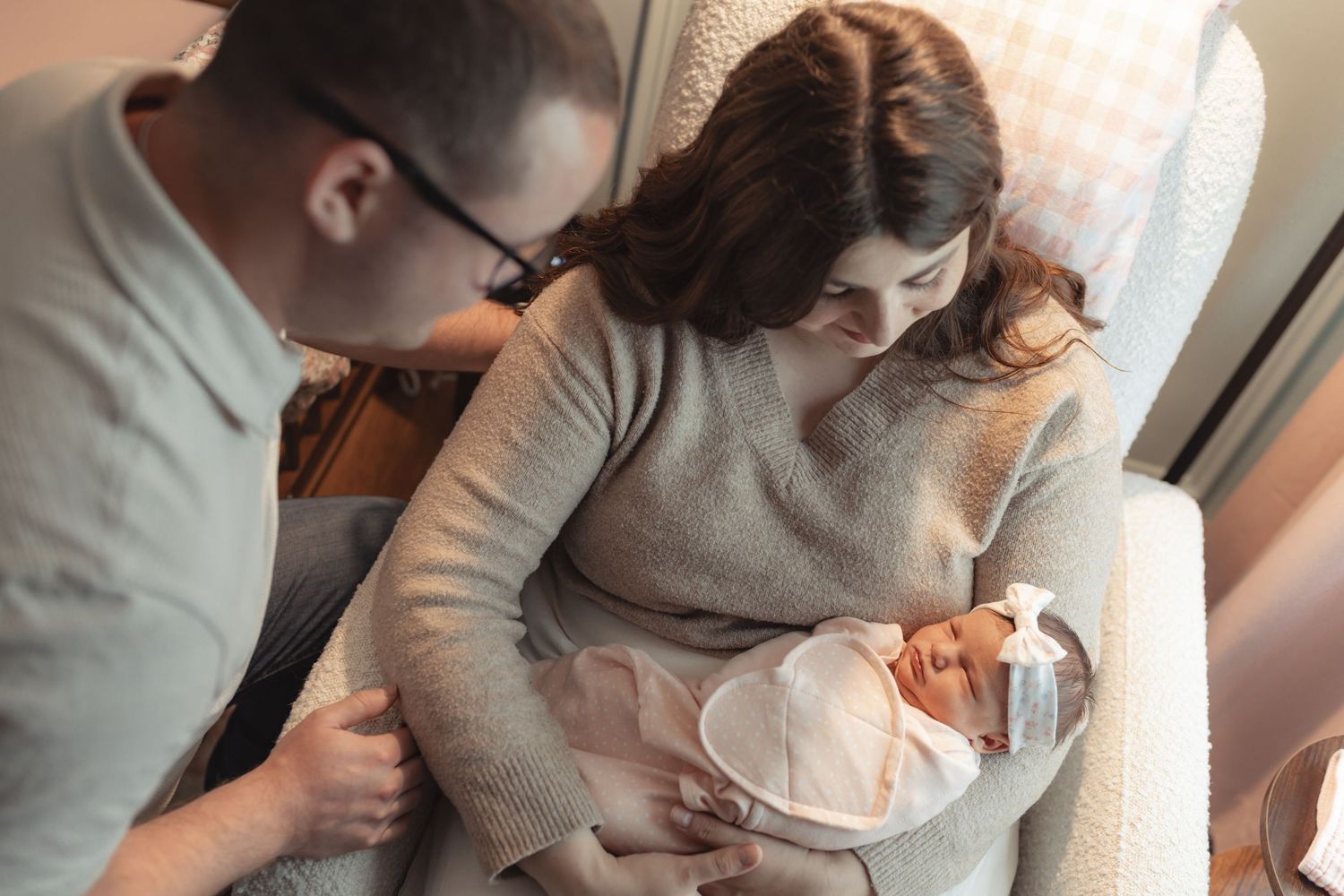 New parents lovingly gaze at their swaddled newborn baby while sitting in a cozy nursery chair.