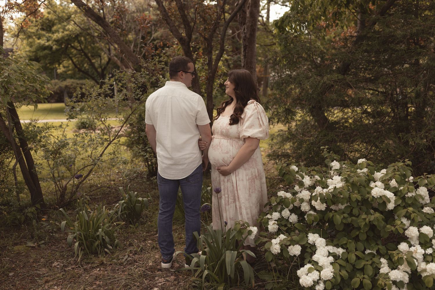 Pregnant couple standing together in a lush garden surrounded by white flowers and green trees.