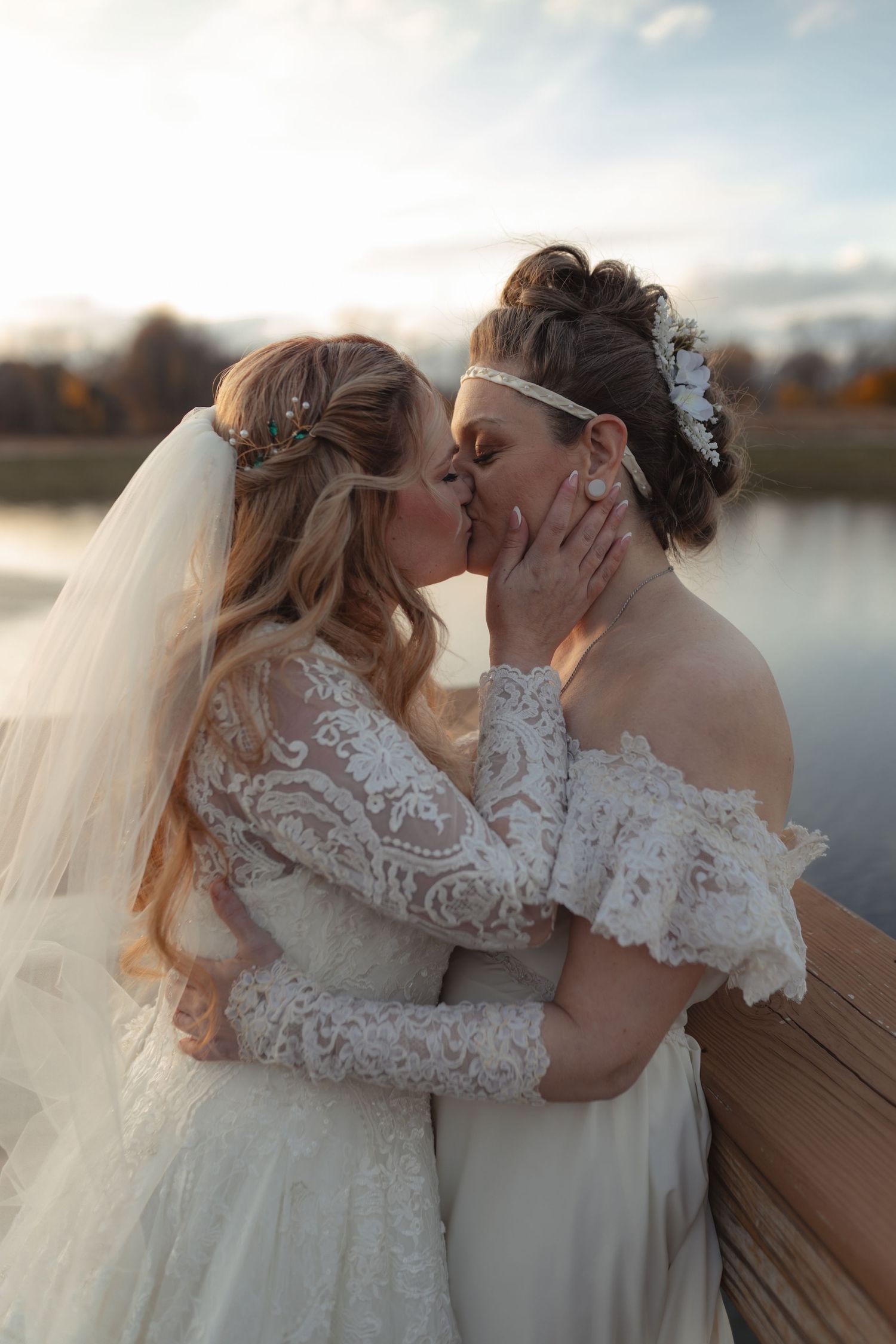 Two brides in elegant lace wedding gowns share a tender kiss at sunset by a serene lake.