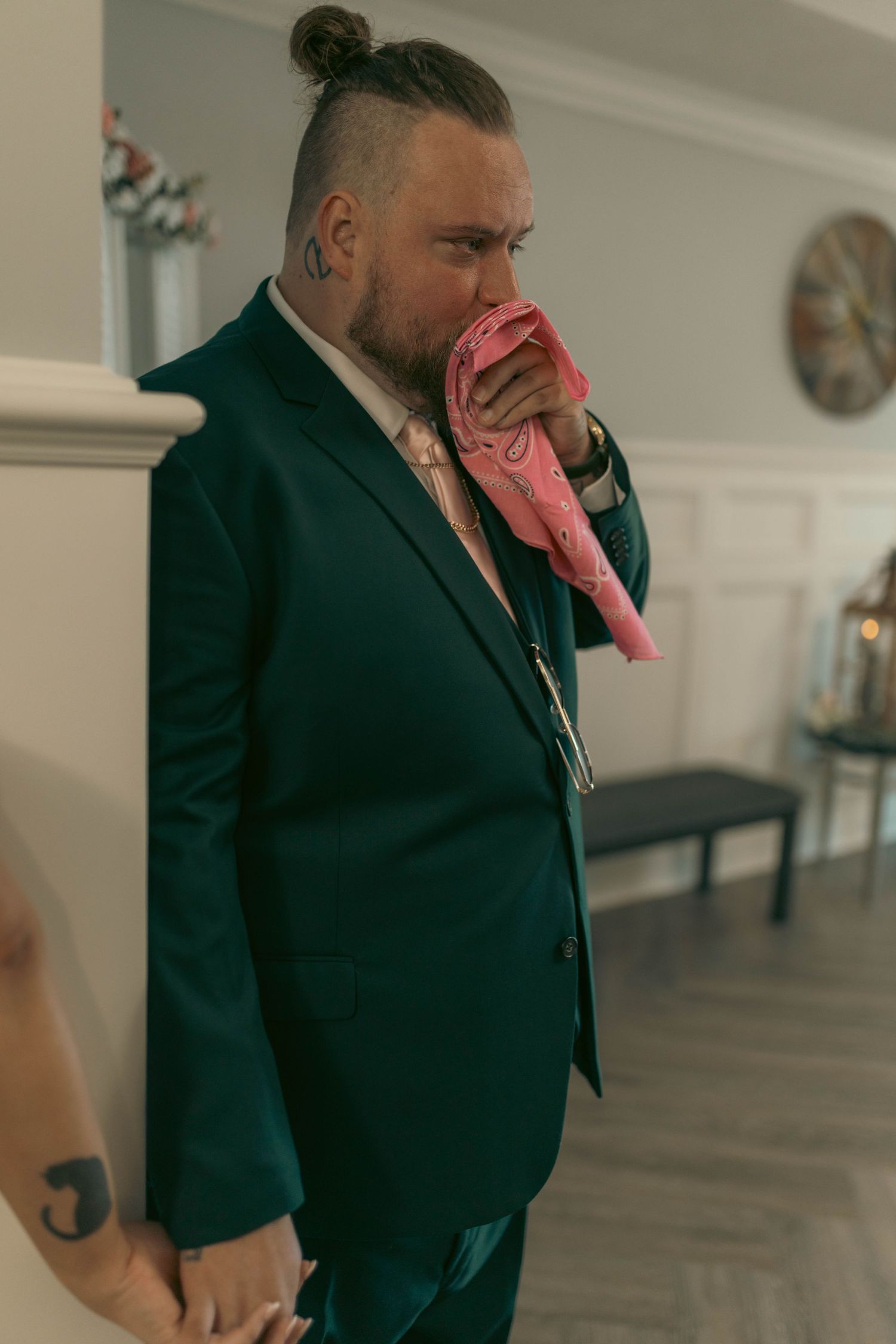 Groom in a dark teal suit kissing a pink item, showcasing face tattoos and hair bun at wedding.