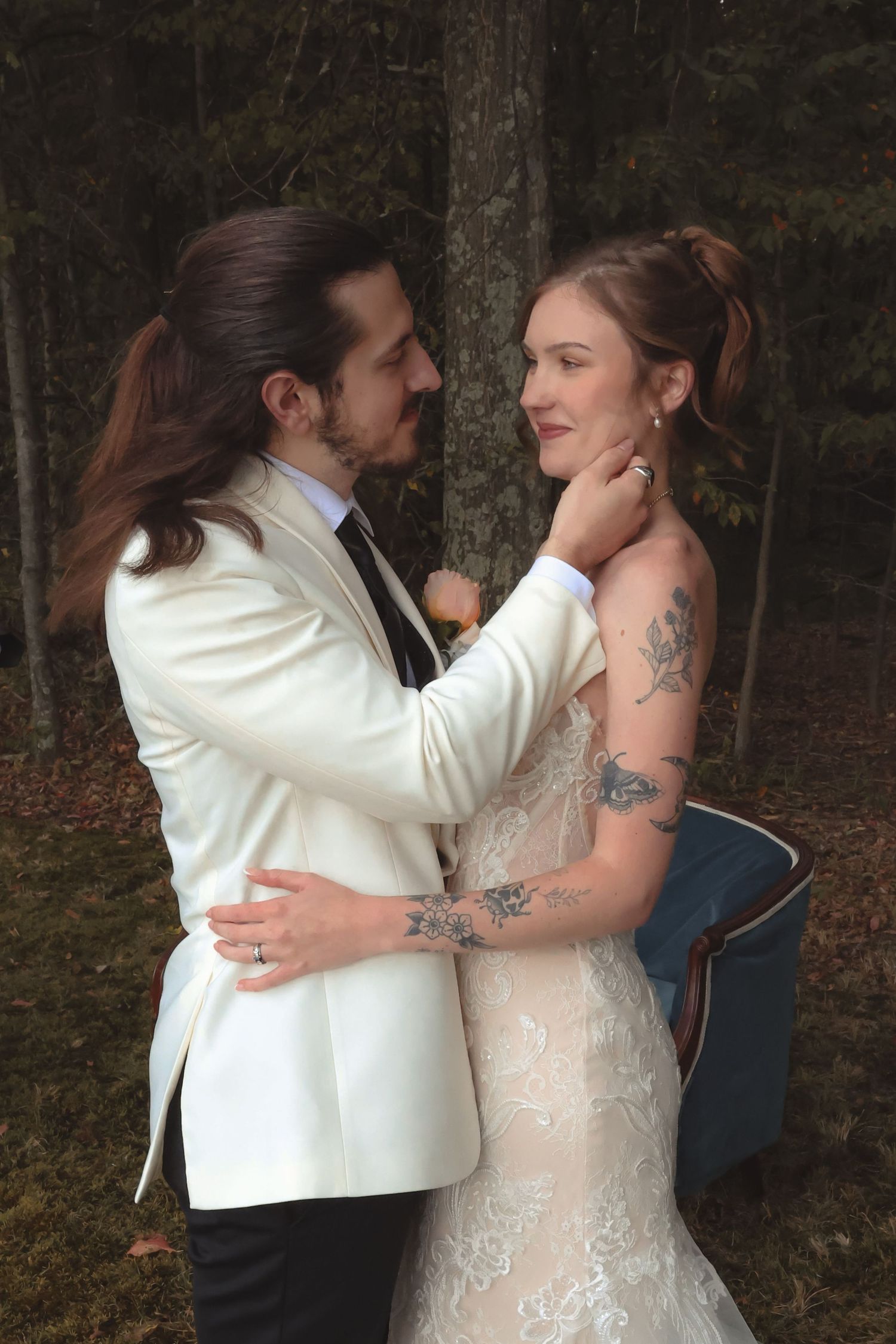 Bride and groom share romantic moment in forest, groom in white tuxedo gently holds brides face.
