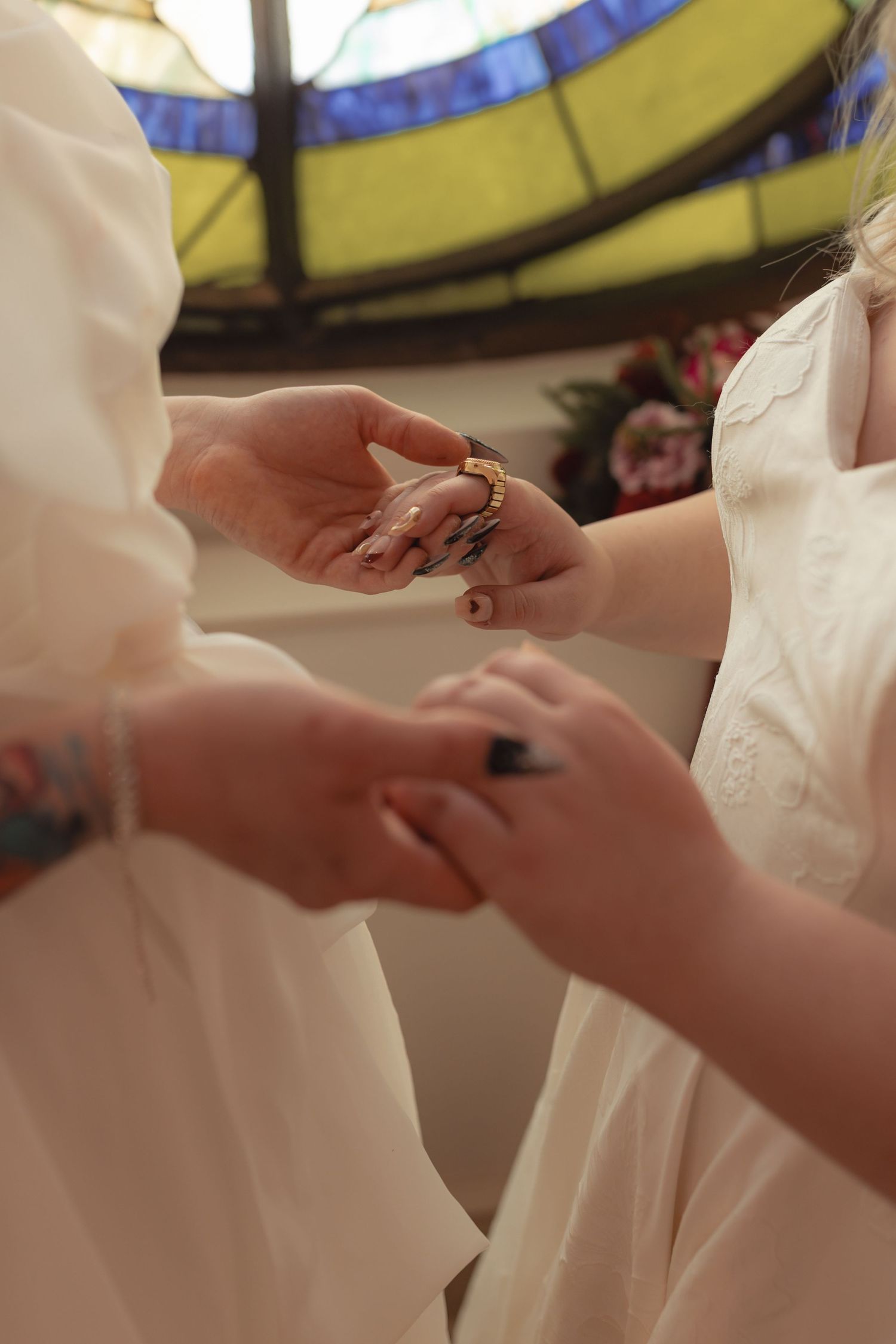 Two brides in white dresses exchange rings during a wedding ceremony beneath a stained glass window.