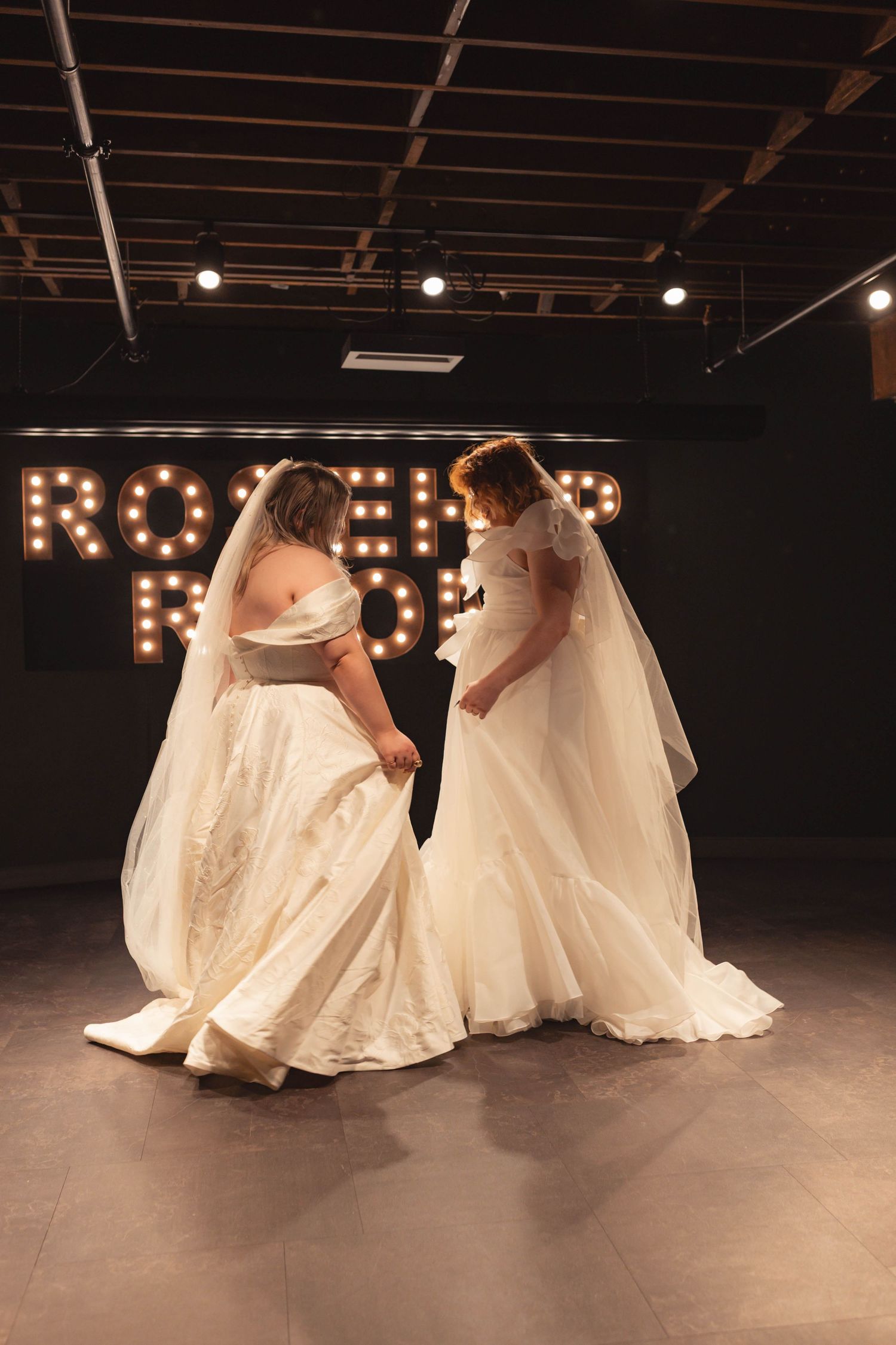 Two brides in white wedding gowns and veils share a tender moment on a dance floor with marquee lights backdrop.