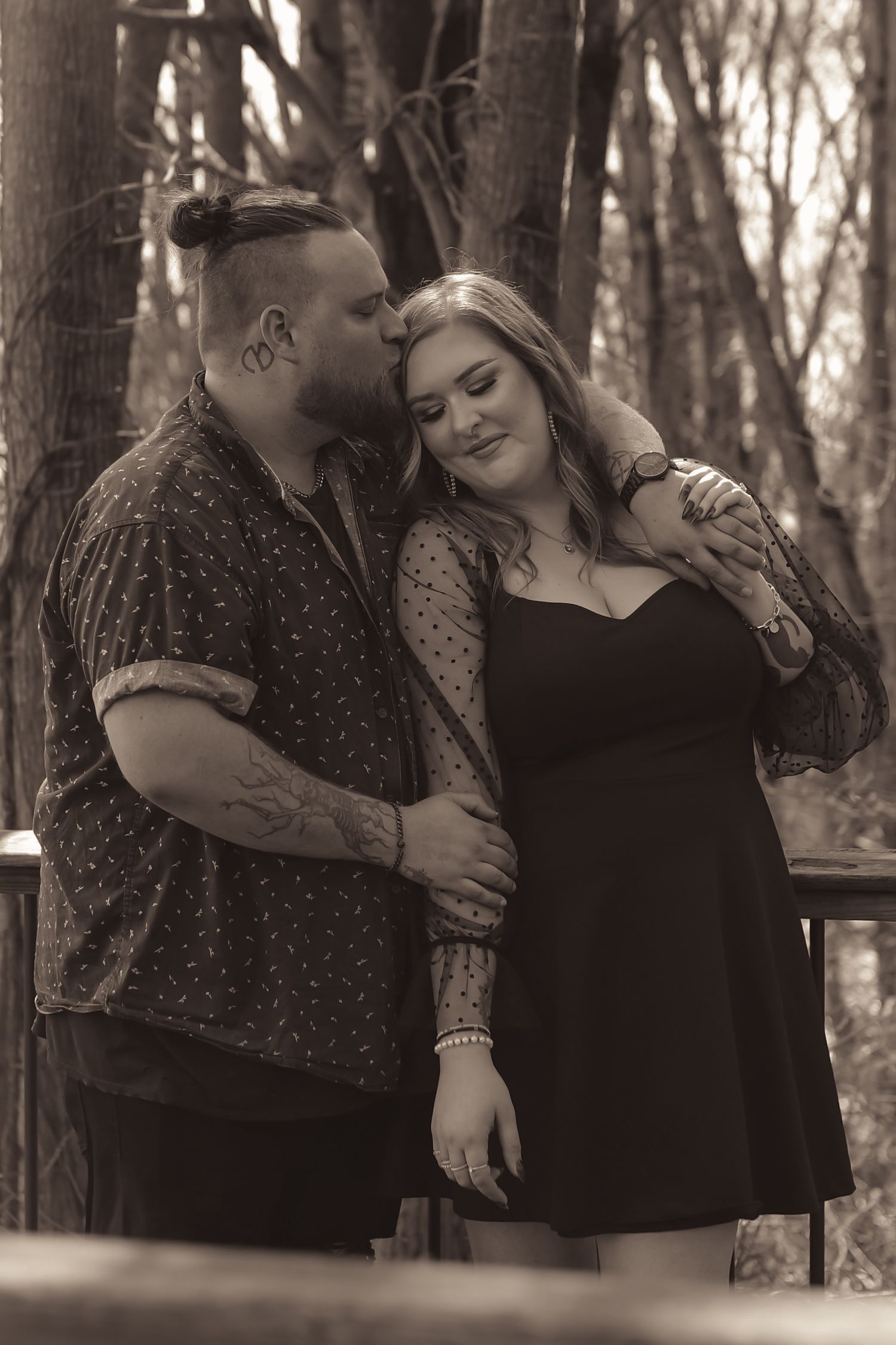 Black and white photo of a tattooed couple embracing outdoors, man kissing womans head in wooded setting.