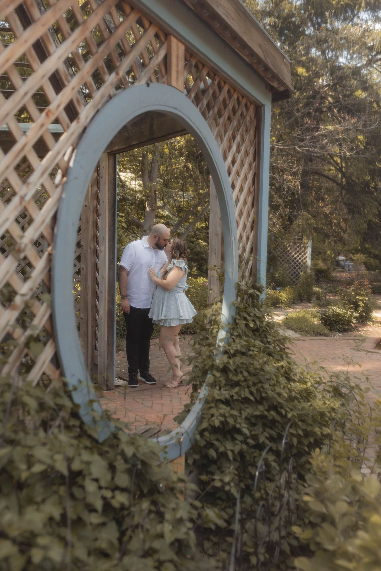 Romantic couple embracing and kissing inside an oval arbor archway surrounded by greenery in a garden.