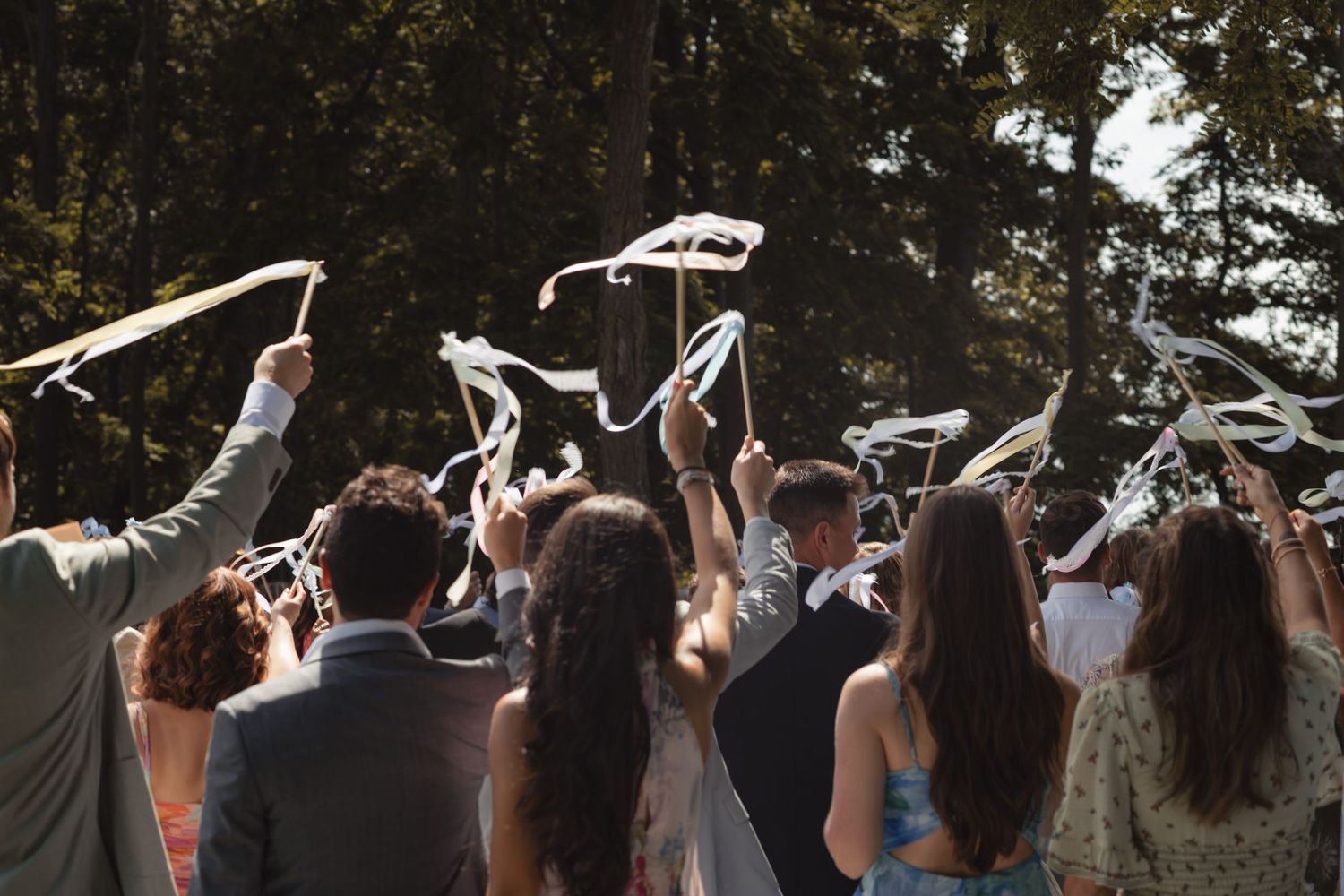 Wedding guests raise ribbon wands in celebration outdoors, surrounded by trees in warm sunlight.