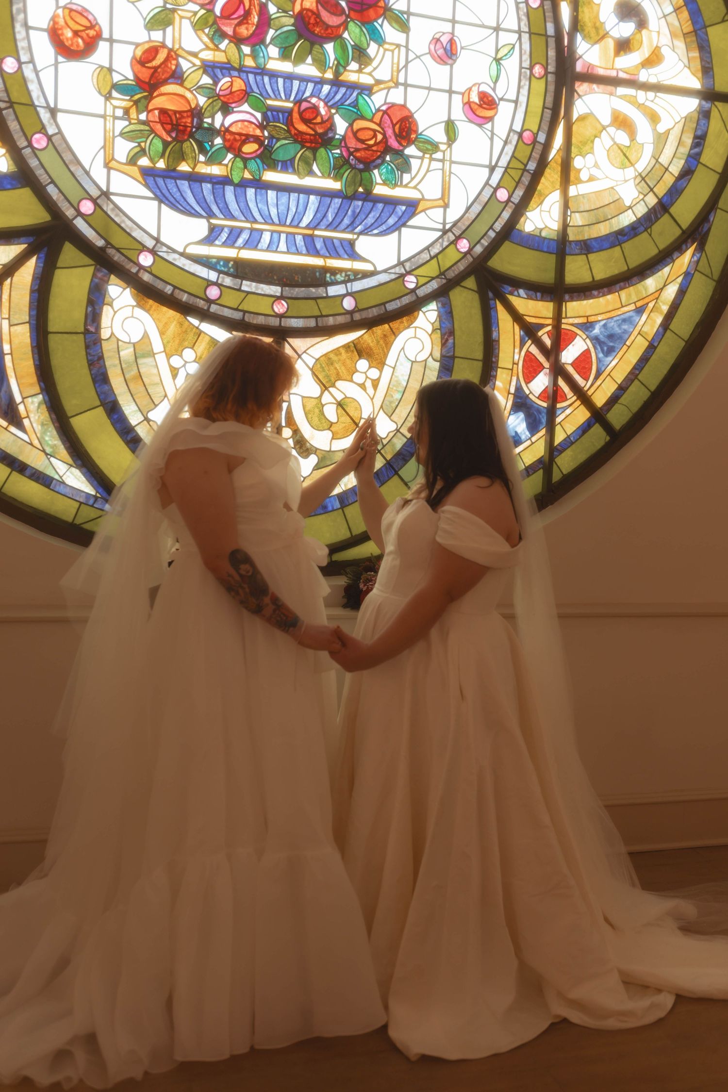Two brides in white gowns holding hands before a stunning circular stained glass window.