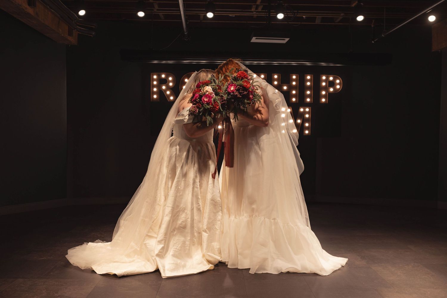 Two brides in white gowns and veils embrace while holding a lush floral bouquet in a softly lit venue.