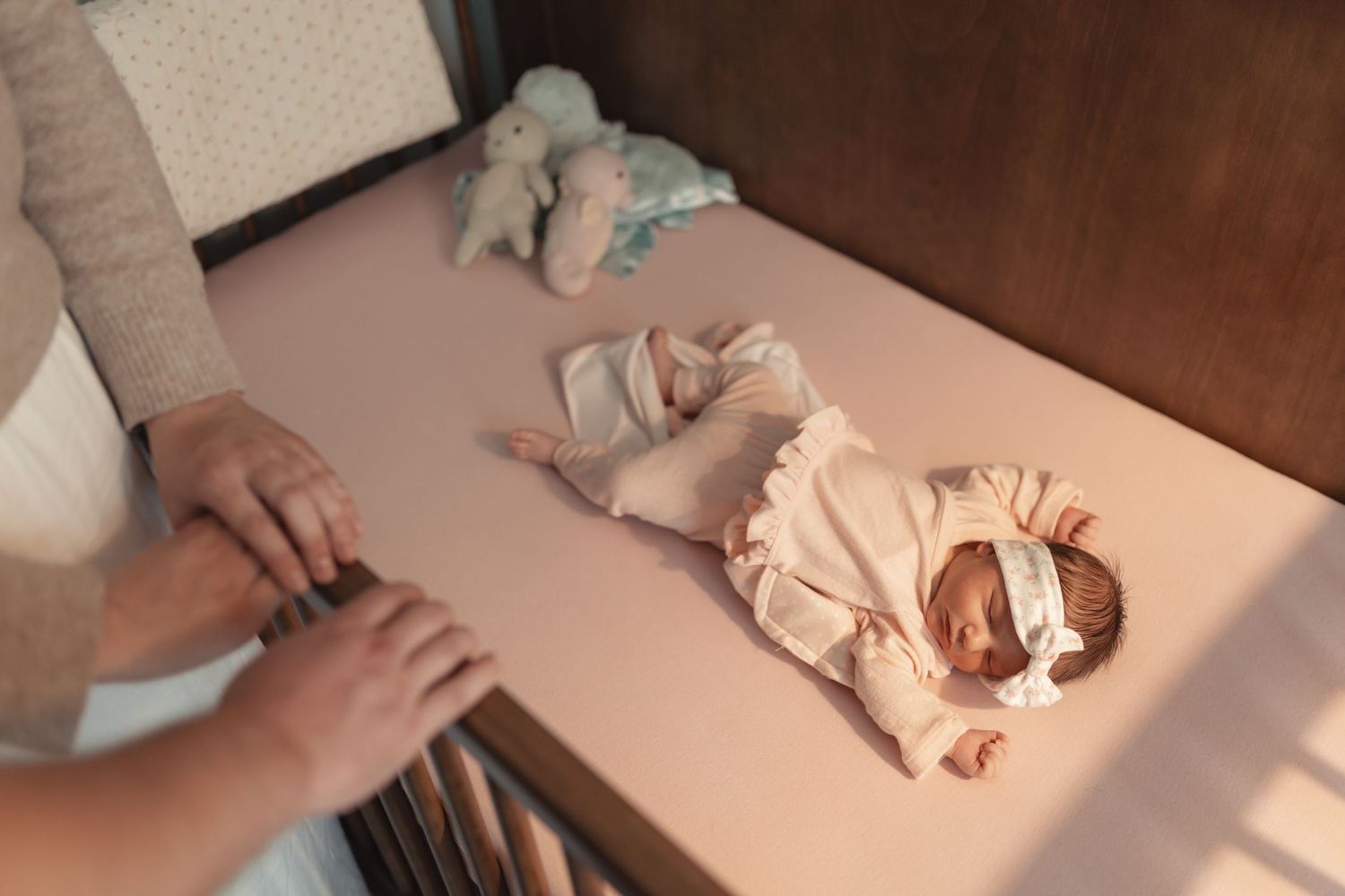 Newborn baby in pink romper and headband lies in a sunlit crib as parent stands nearby.