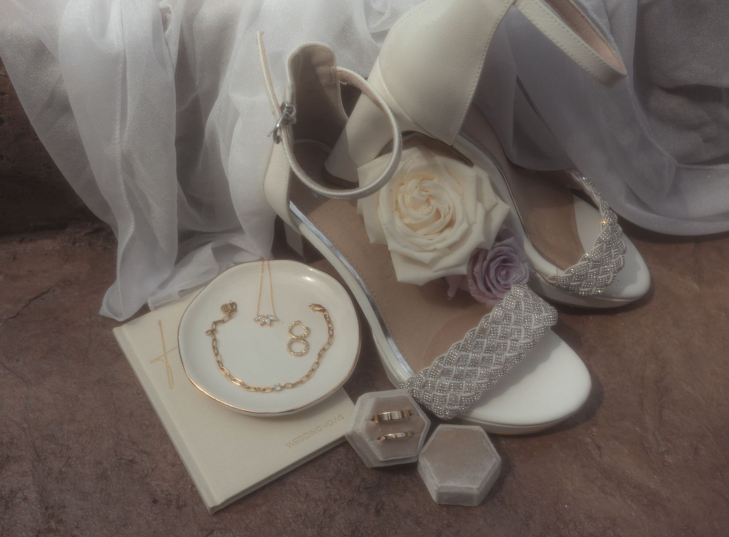 Elegant bridal accessories featuring white heeled sandals, floral decor, gold necklace on ring dish, and diamond earrings in gray box.