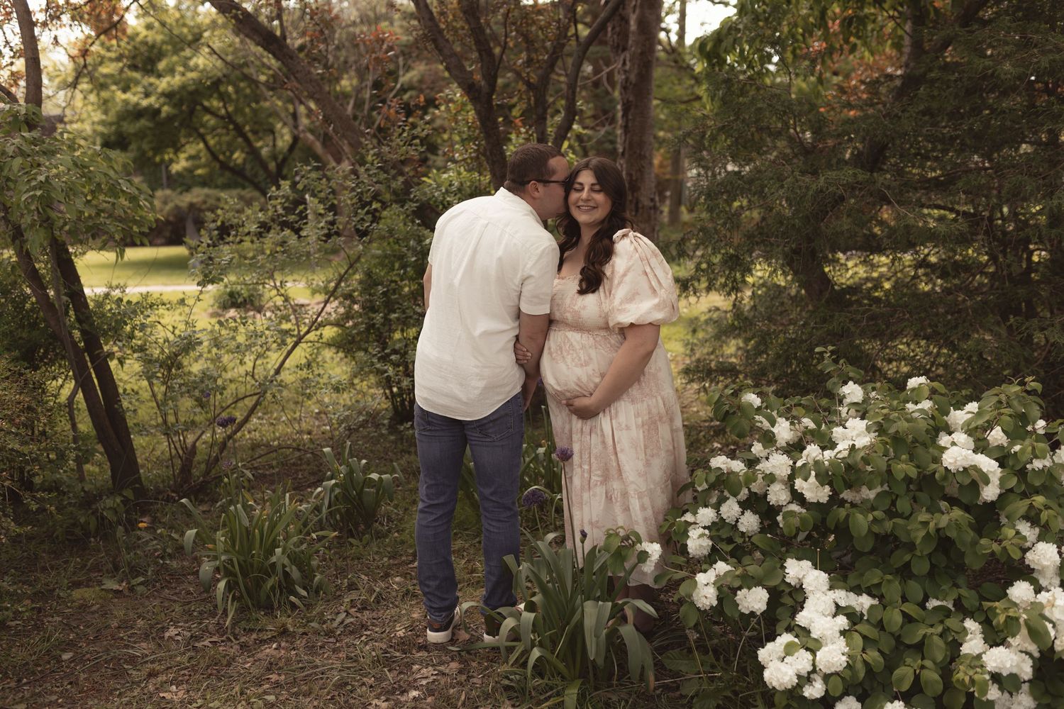 Couple sharing a tender moment in a lush garden, woman in flowy dress cradling baby bump during maternity photoshoot.
