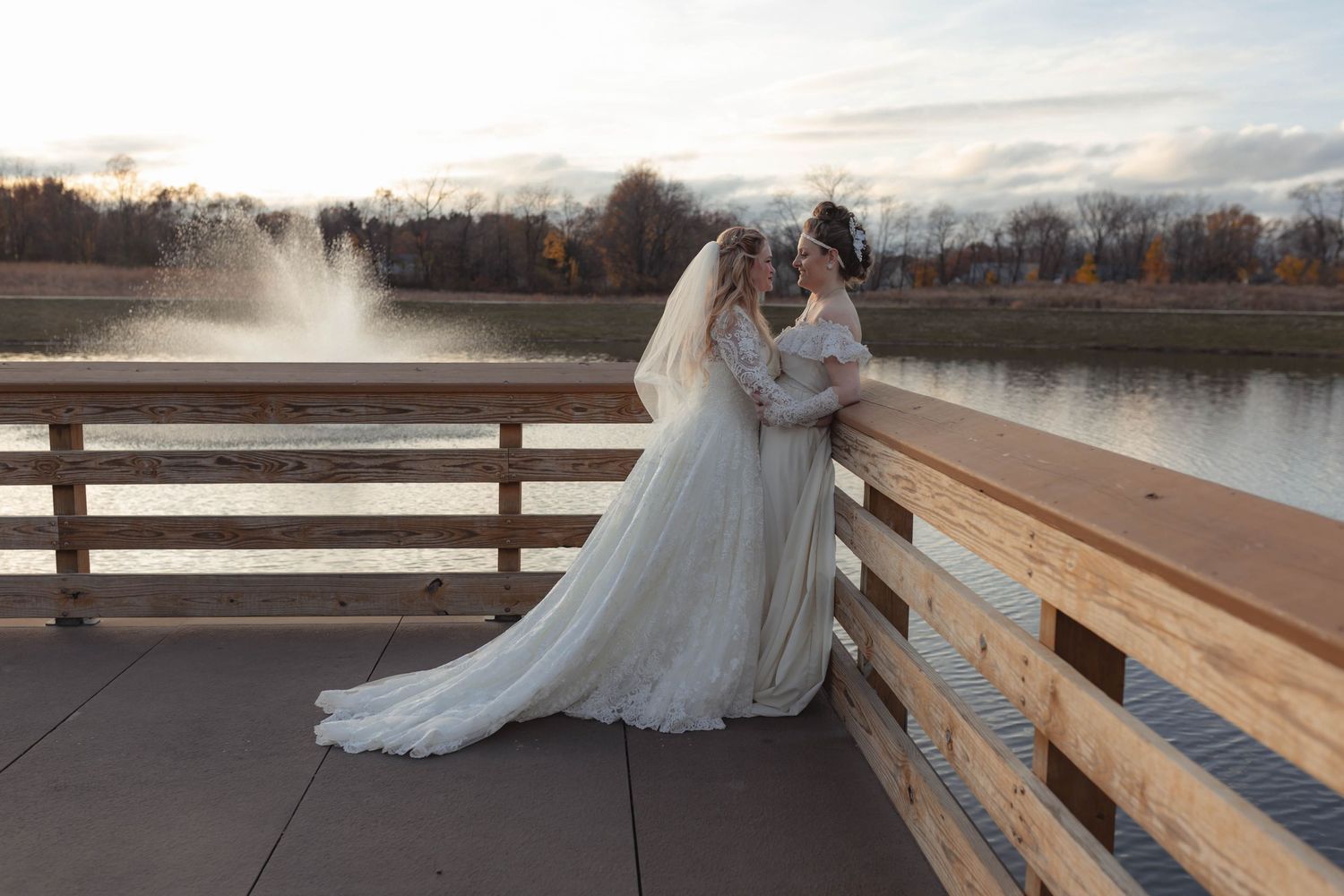 Two brides in elegant white gowns embrace on a wooden bridge with a fountain and autumn trees in the background.