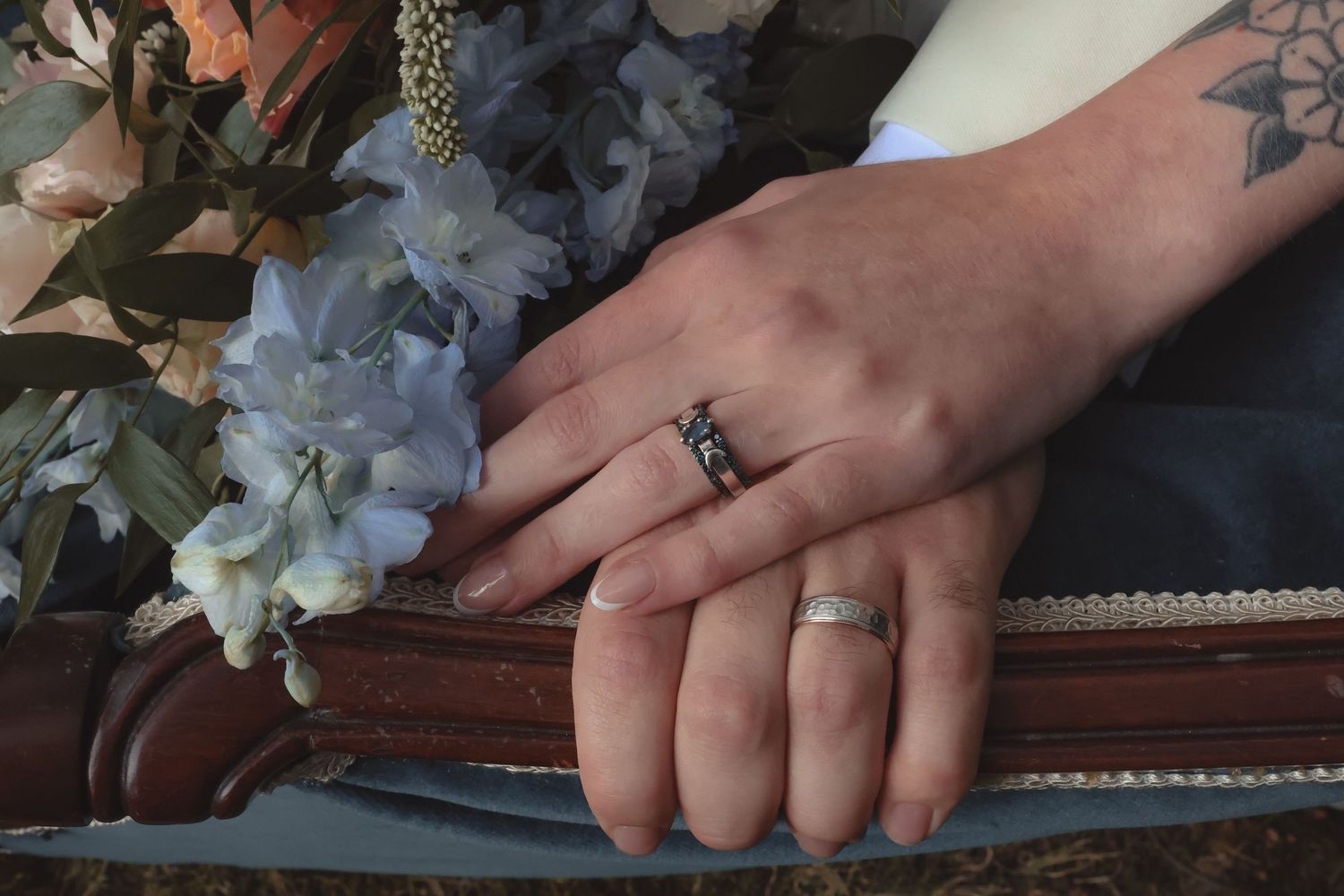 Two hands with wedding rings resting on a surface beside blue hydrangeas, one hand featuring a delicate tattoo.