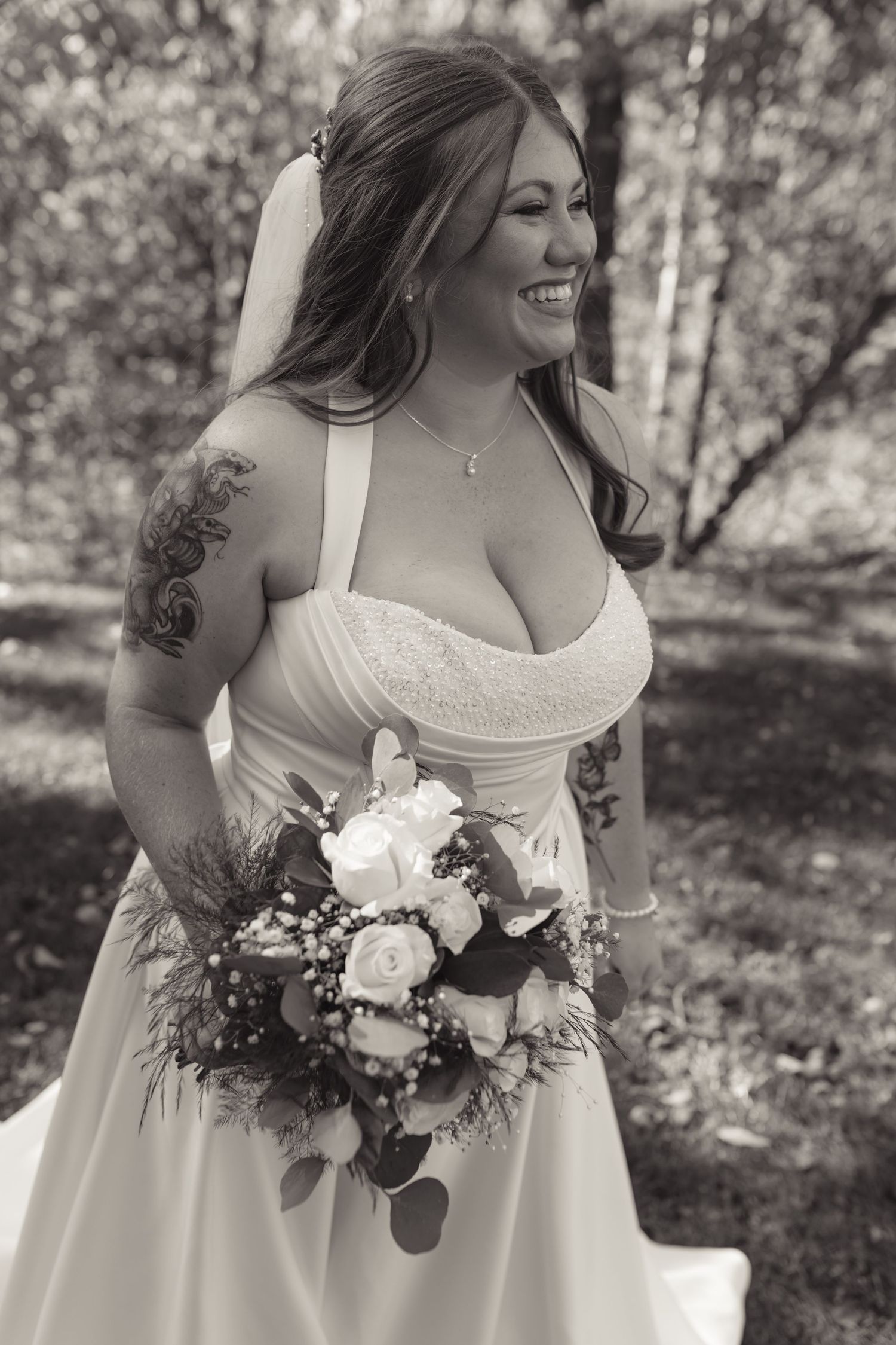 Smiling bride in white gown holding floral bouquet with roses, wearing veil and floral arm tattoo, black and white outdoor photo.
