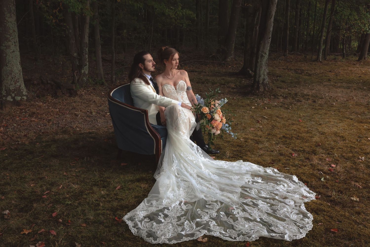 Bride in lace gown with long train sits with groom in forest clearing holding a bouquet of flowers.