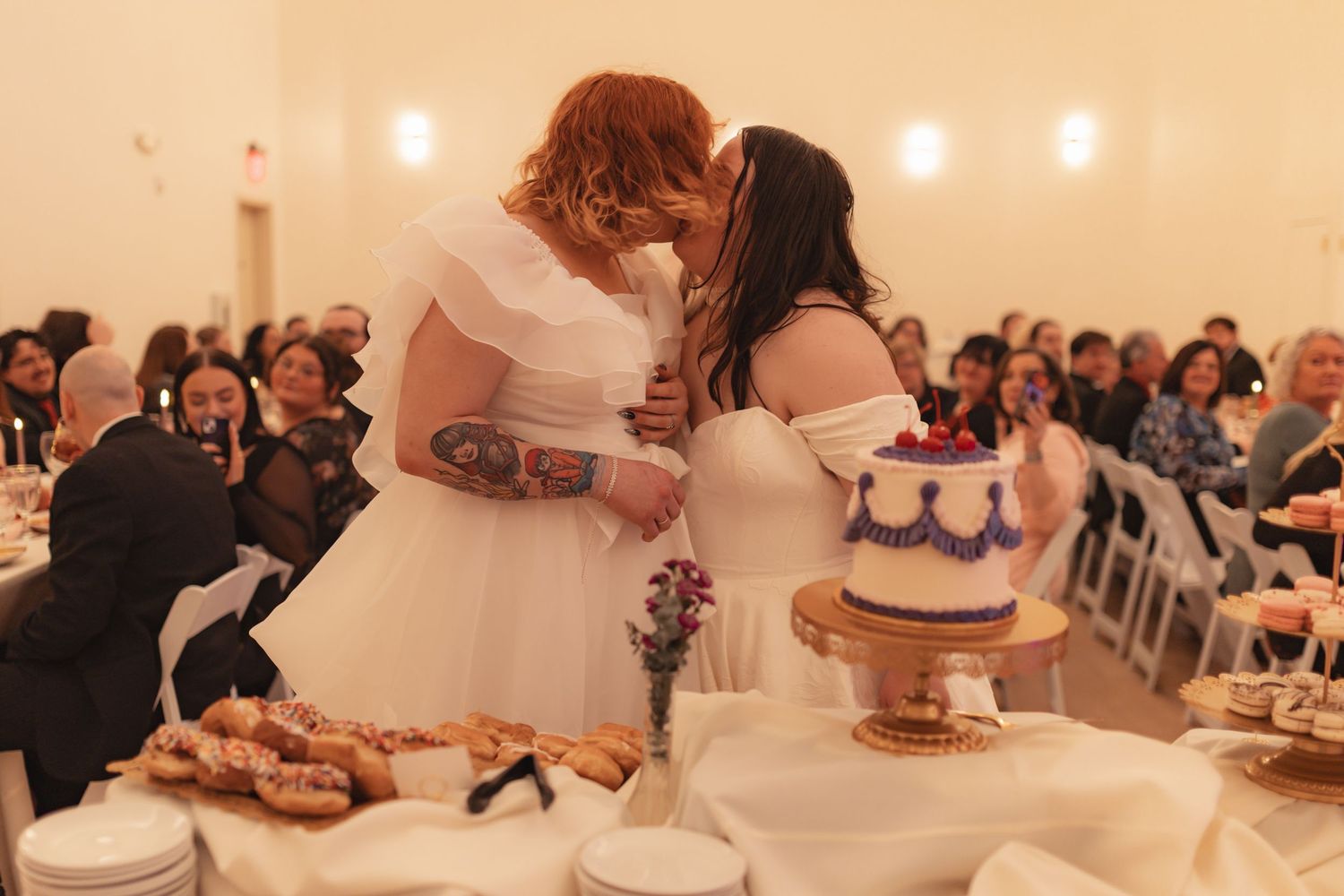 Two brides in white dresses share a kiss at their wedding reception beside a decorated cake.