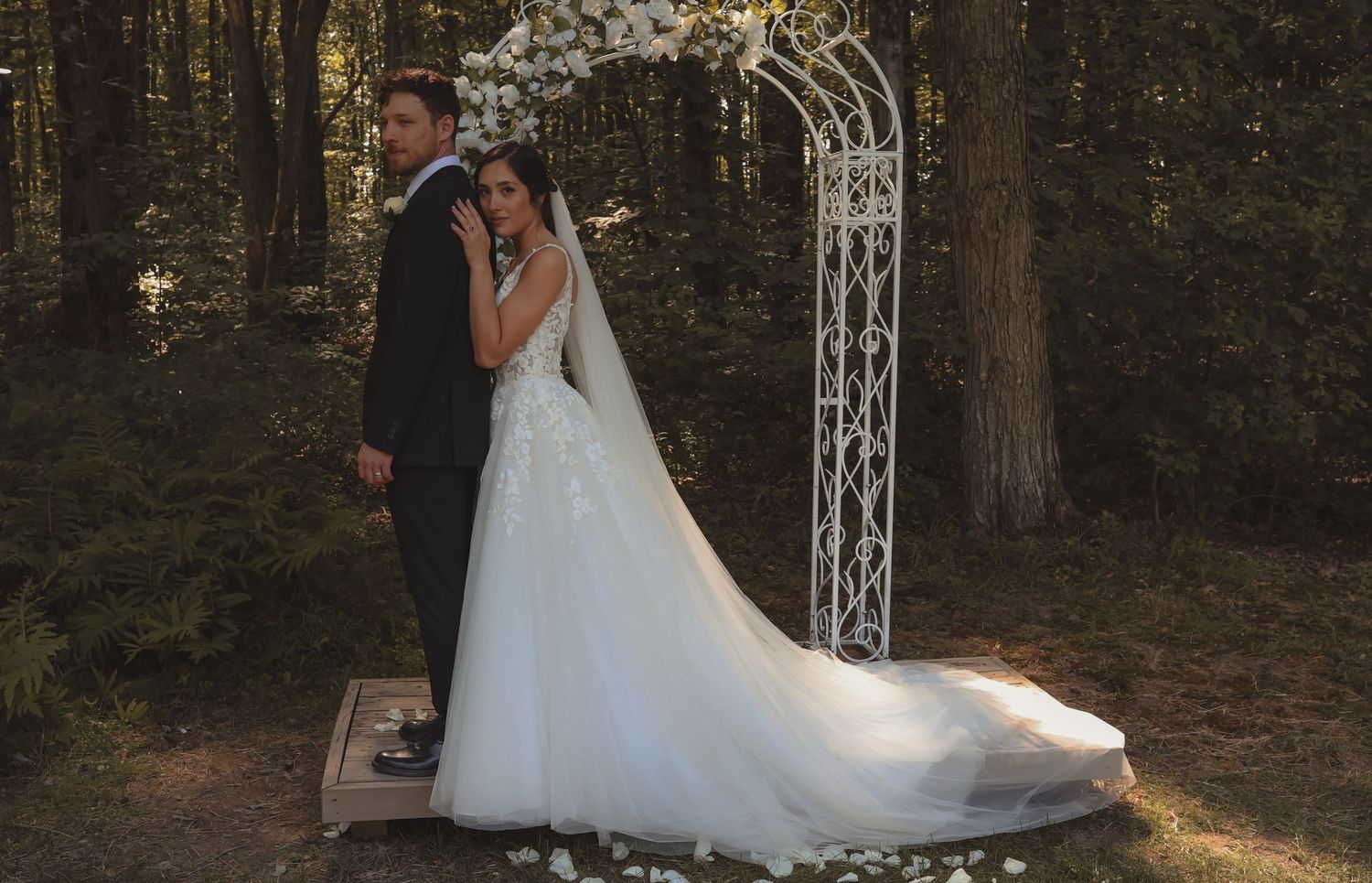 A bride and groom pose together under a floral arch in a forest wedding ceremony setting.
