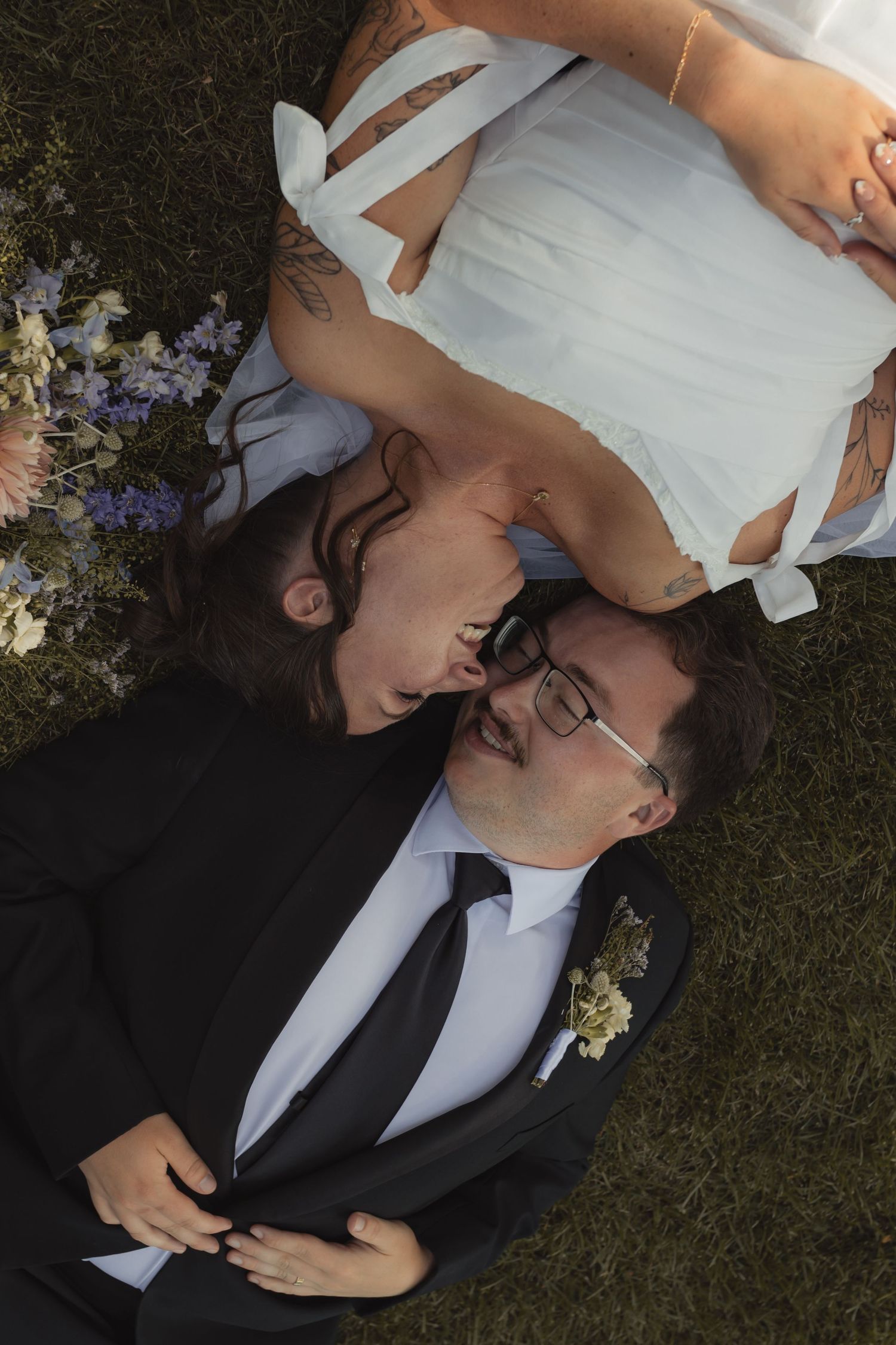 Bride and groom lying on grass laughing together, wearing wedding attire with bouquet nearby, aerial view.