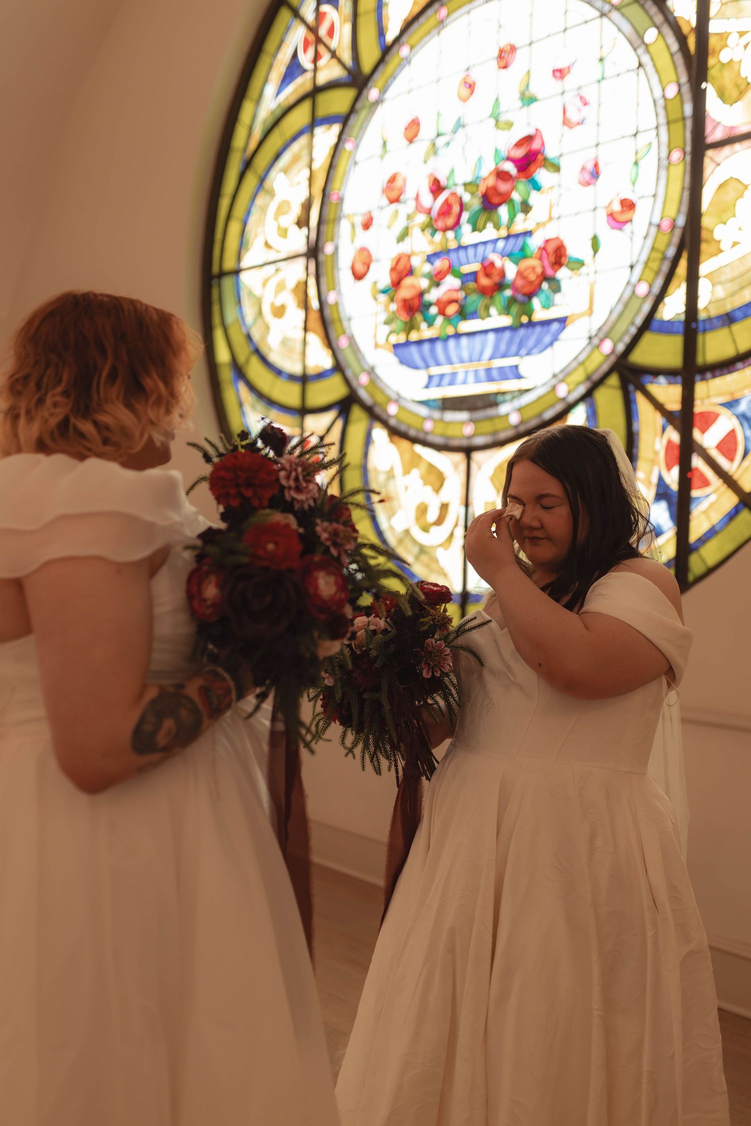 Two brides in white dresses share an emotional moment before a stained glass rose window.