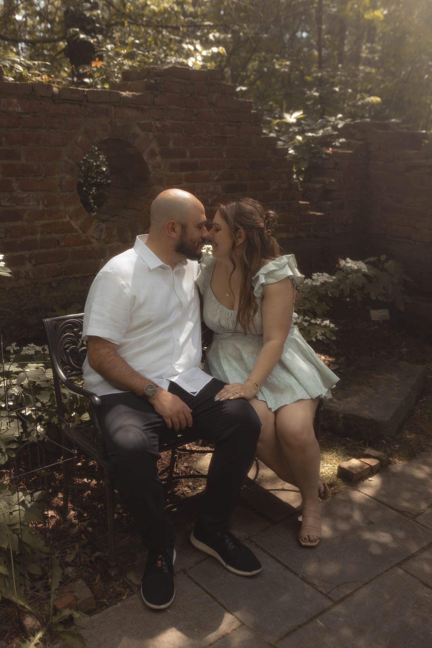 Couple sharing a romantic moment on a garden bench surrounded by lush greenery and rustic brick wall.