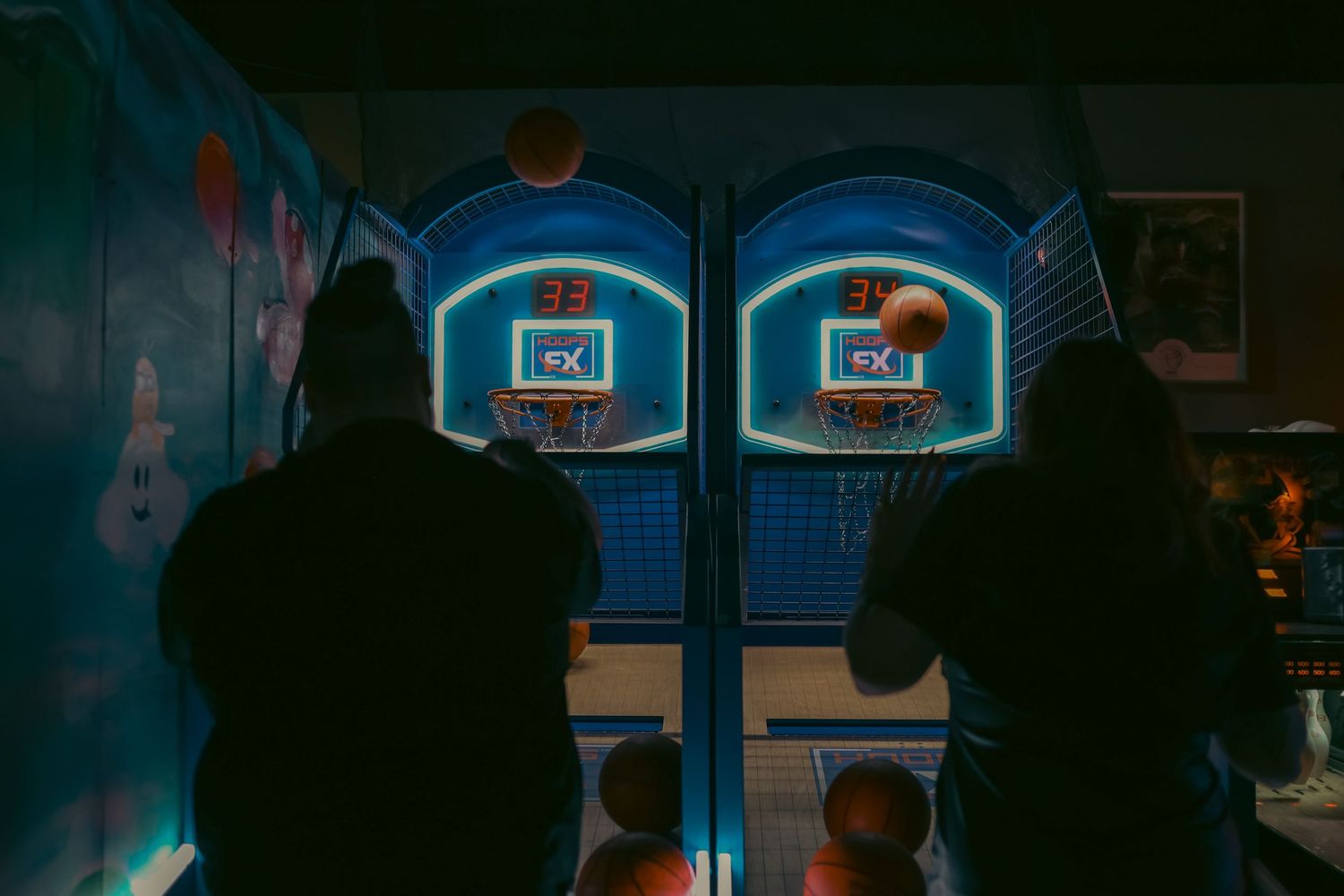 Two people playing arcade basketball game in dark neon-lit room.