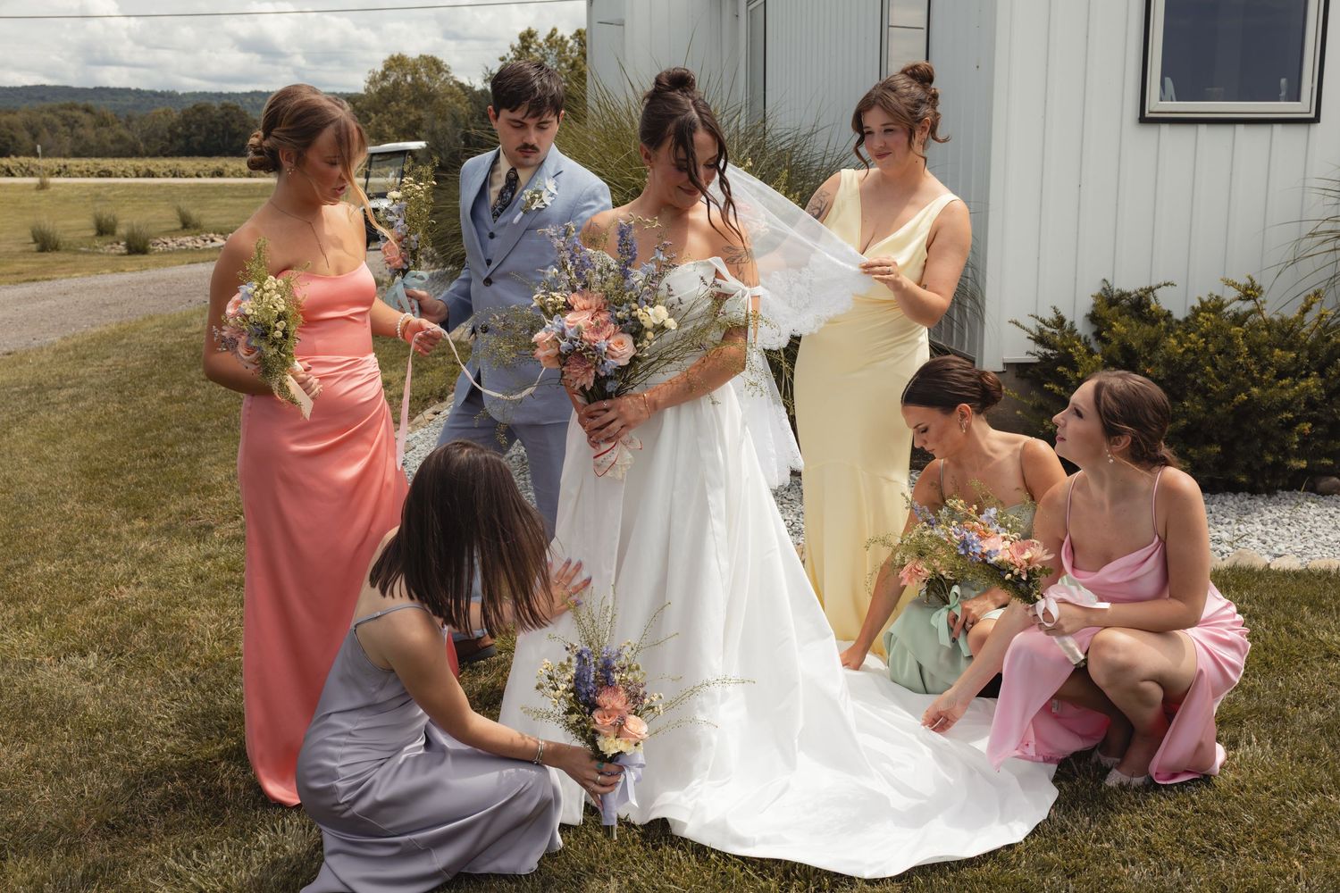 Bride in white gown surrounded by colorful bridesmaids adjusting her dress outdoors on a sunny wedding day.