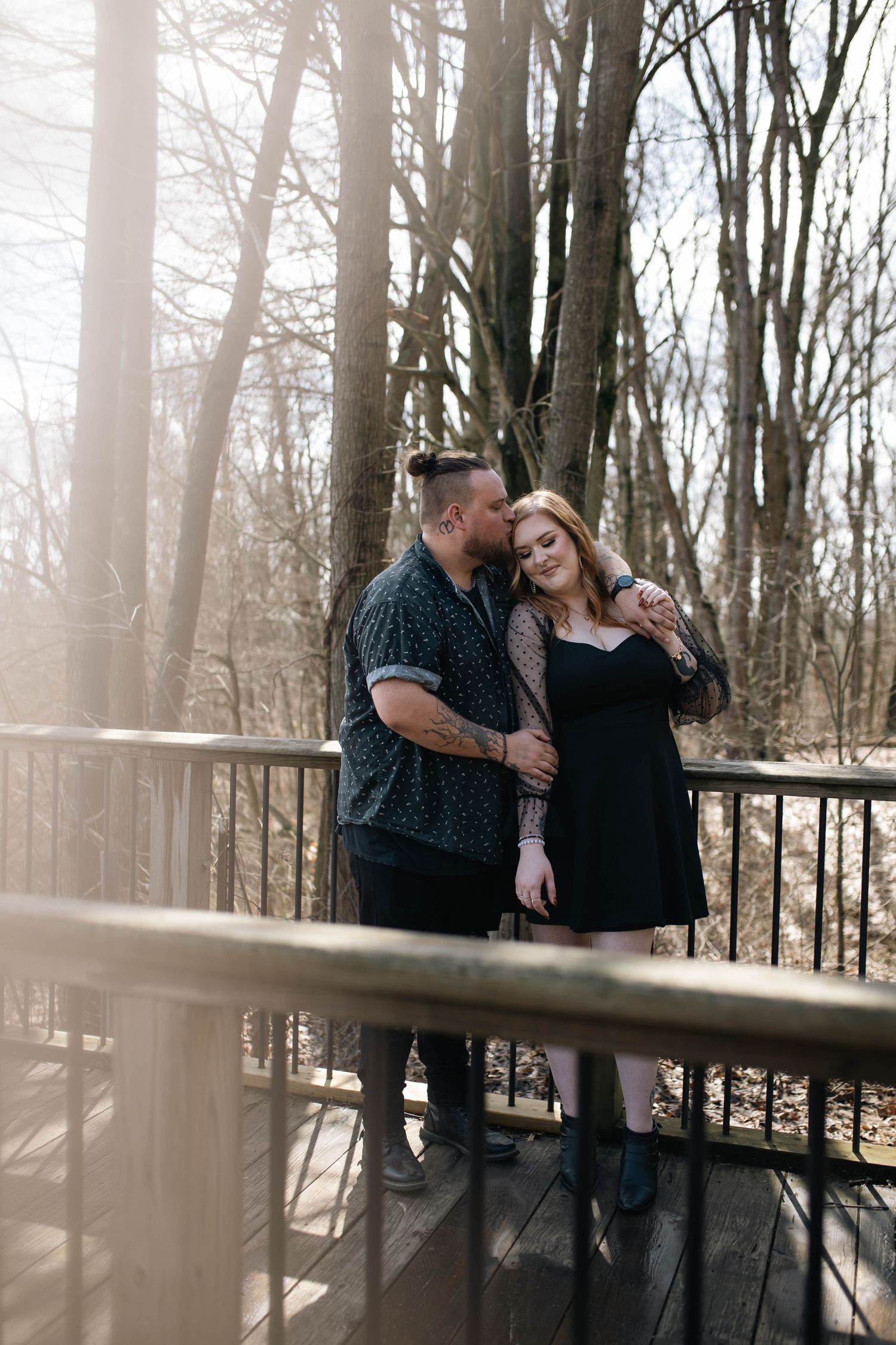 Couple embracing on a wooden bridge surrounded by bare trees in soft sunlight.