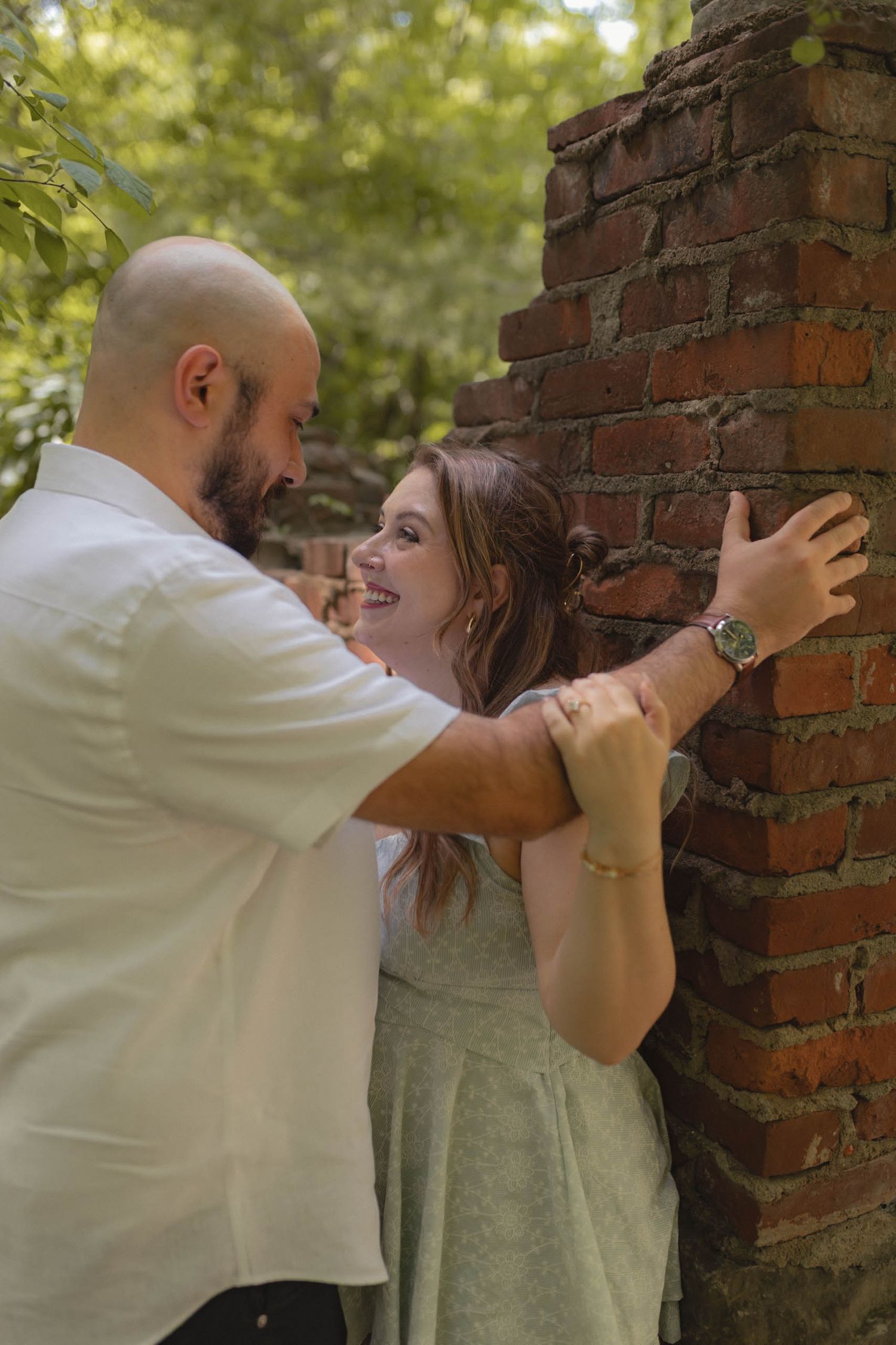 Couple sharing a romantic moment against a rustic brick wall surrounded by lush greenery.
