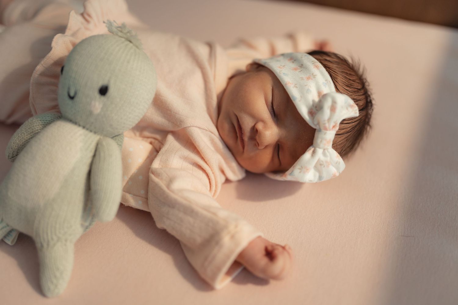 Newborn baby girl sleeping peacefully in crib wearing pink outfit and white bow headband beside mint green bunny toy.