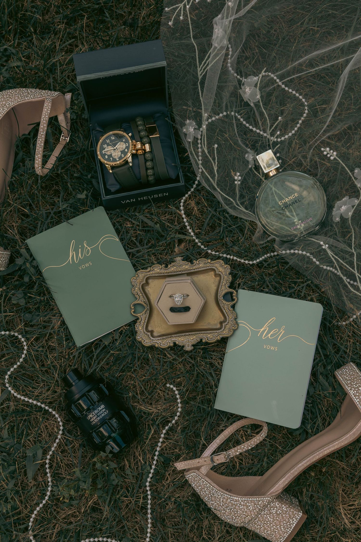 Wedding flat lay featuring his and her vow books, rings, watch, heels, pearls, and perfume on grass.