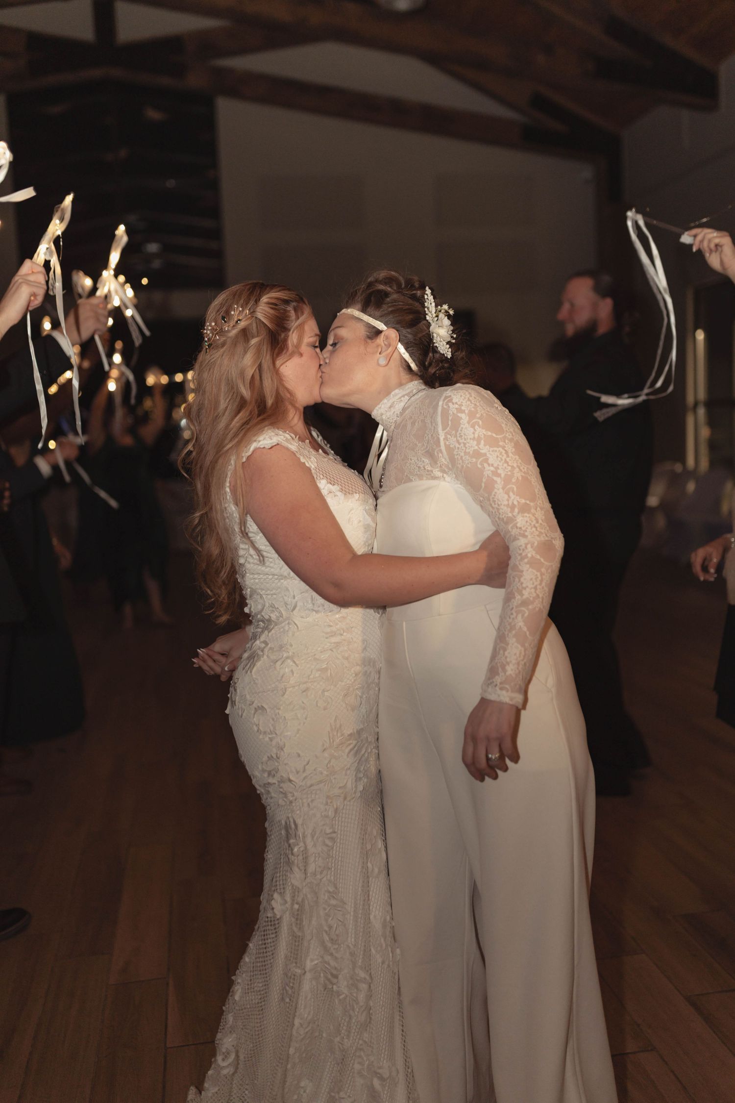 Two brides sharing a first dance kiss surrounded by guests with sparklers at wedding reception.