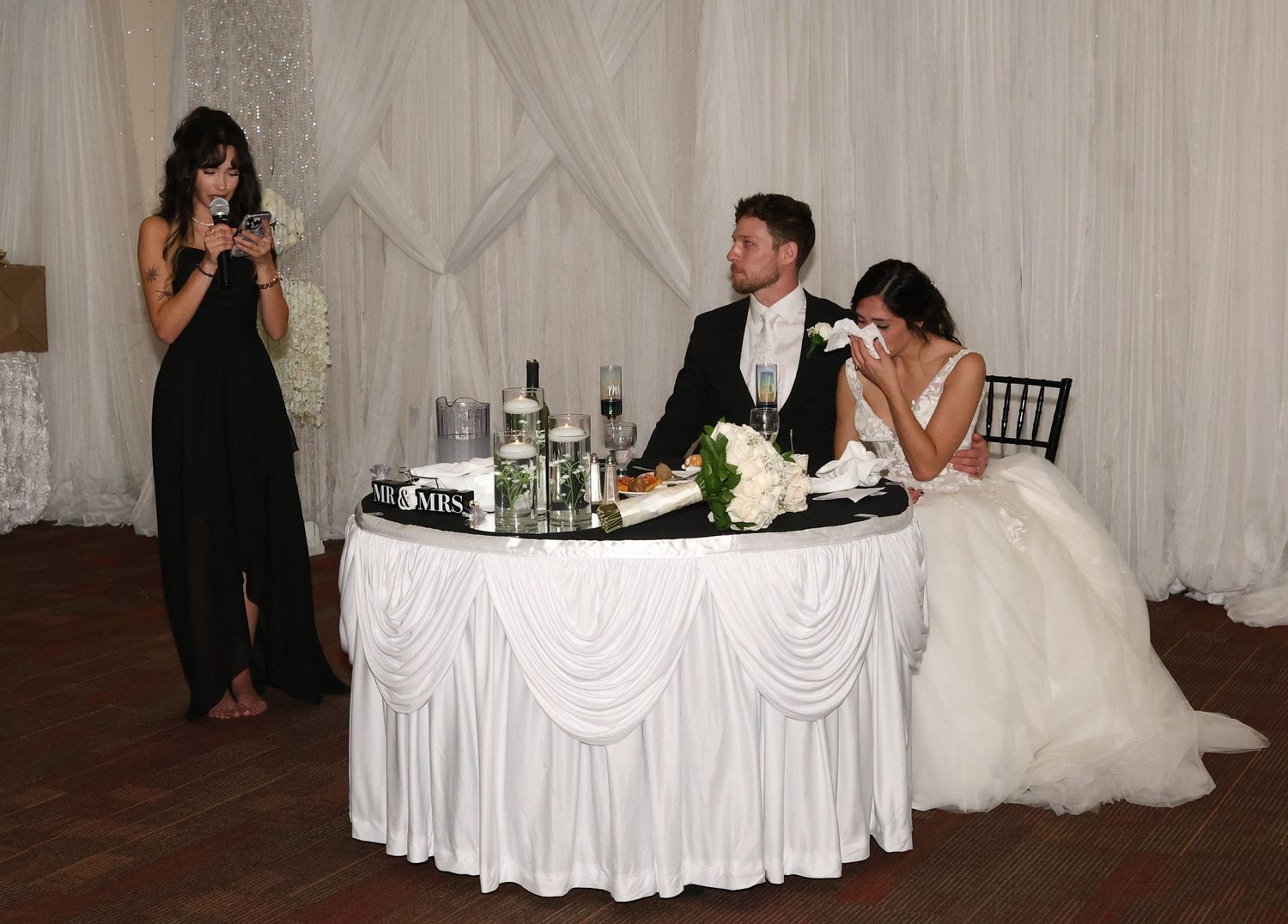 Bride and groom seated at sweetheart table while bridesmaid delivers emotional wedding toast at elegant reception.