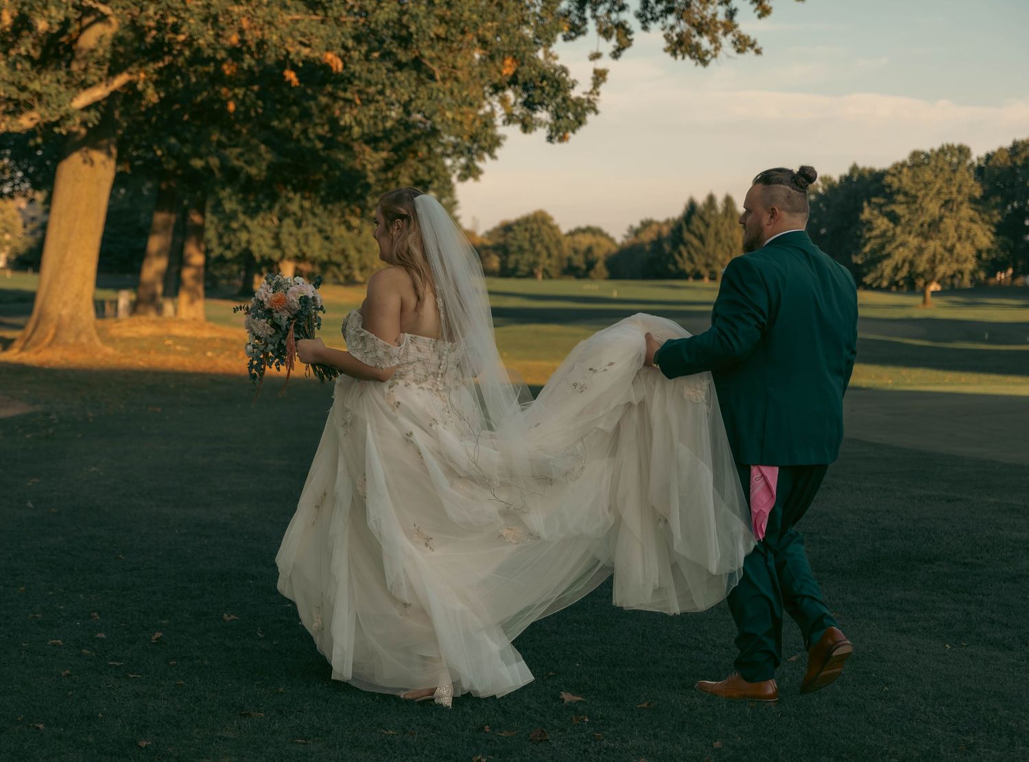 Bride in flowing white gown and veil walks with groom in green suit across park at golden hour.