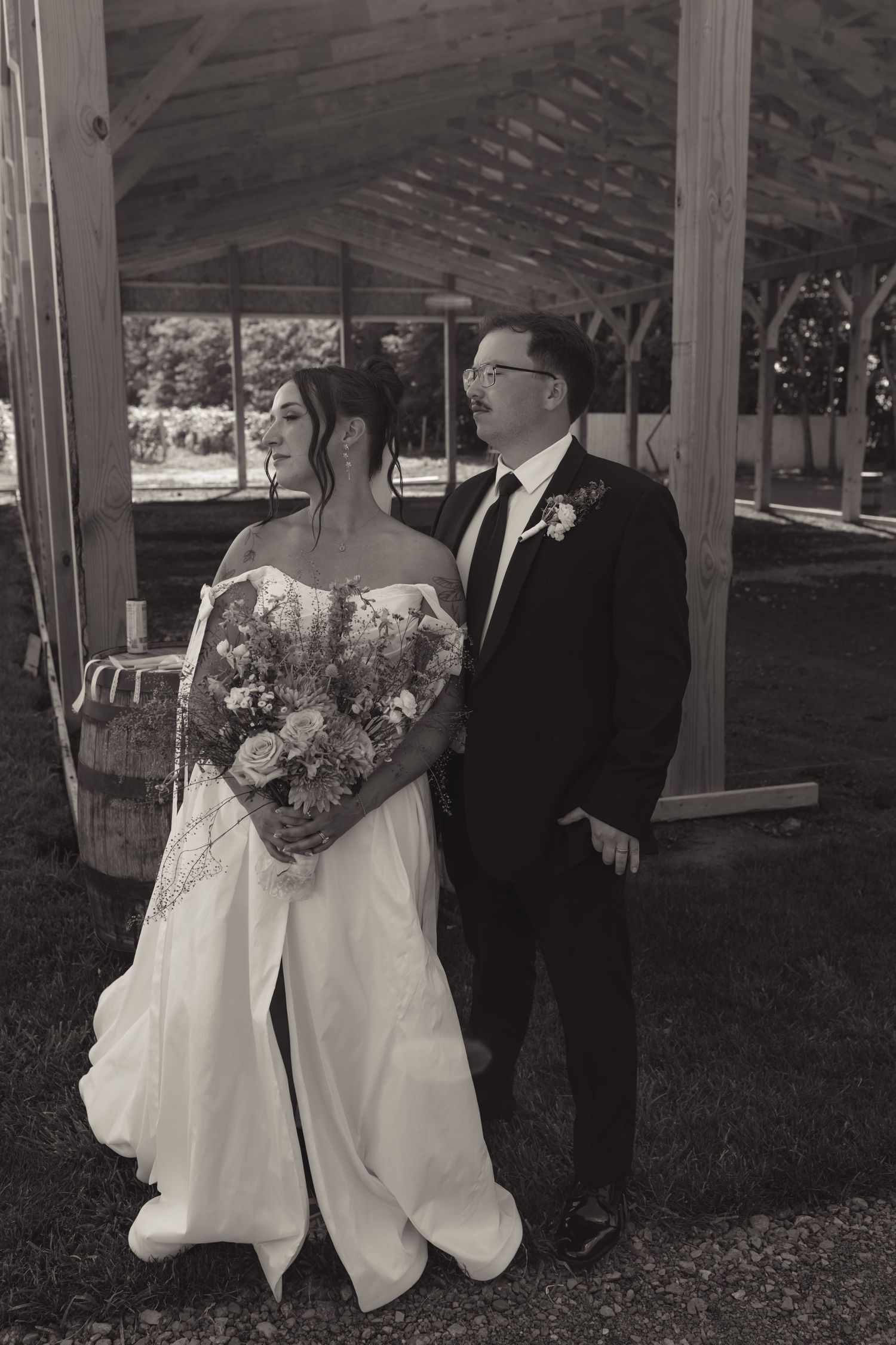Black and white photo of a bride and groom standing together outdoors under a wooden pergola on their wedding day.