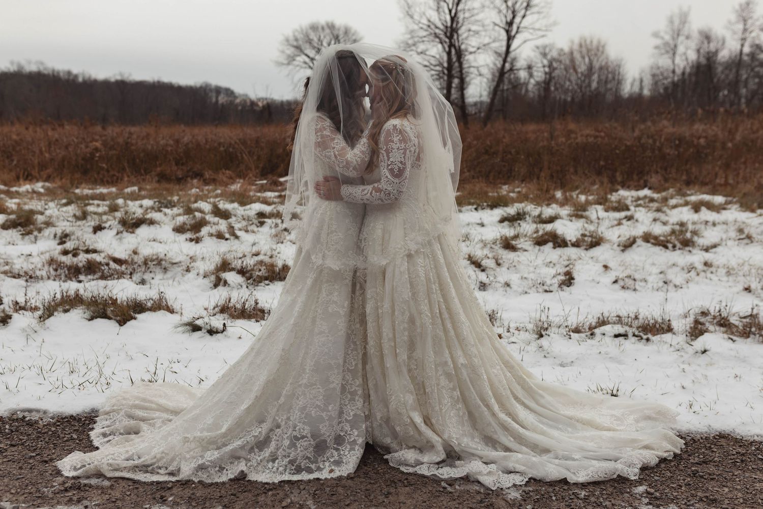 Two brides in white lace gowns and veils share an intimate moment in a snowy winter field.