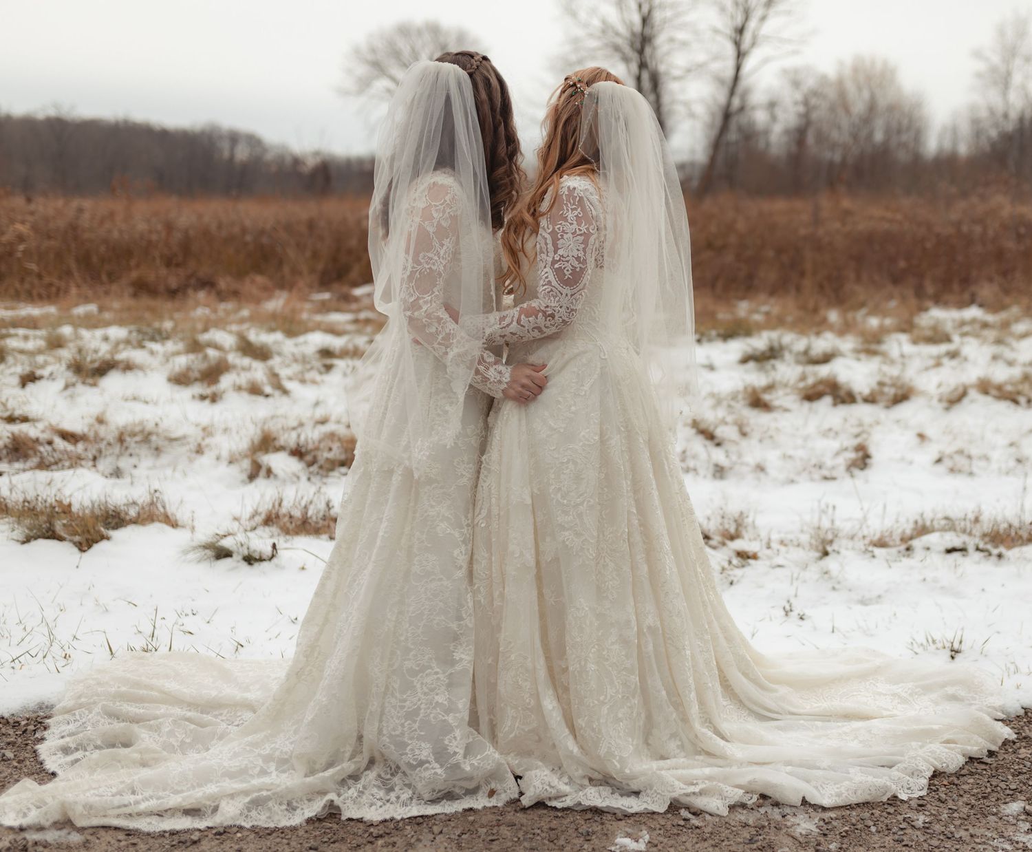 Two brides in lace wedding dresses with veils standing back-to-back in snowy winter field.
