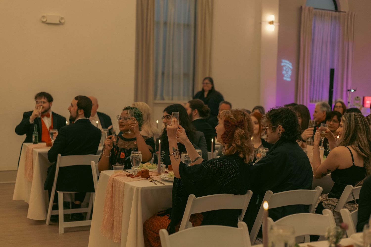 Elegantly dressed wedding guests seated at white-draped tables during a formal reception dinner in a softly lit venue.